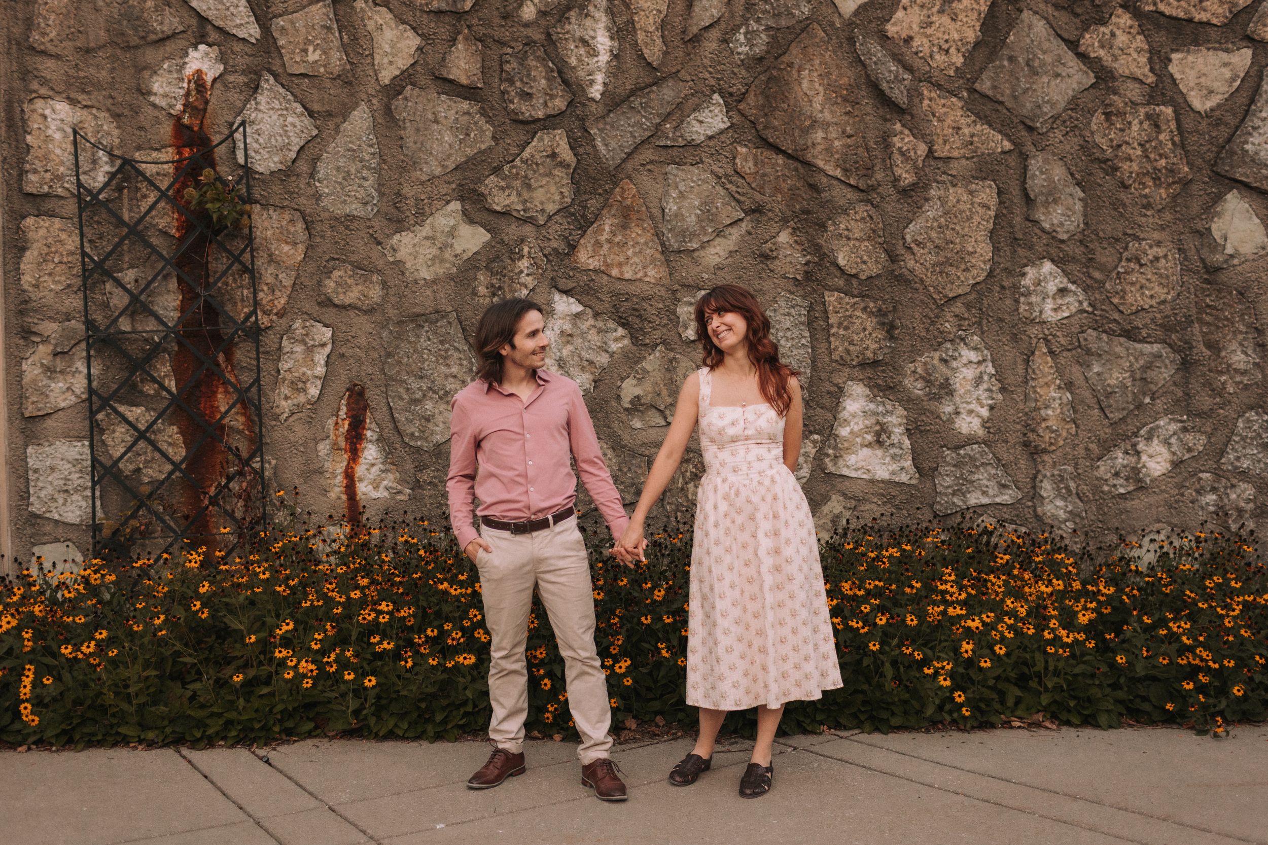 A couple poses against a stone wall with blooming orange flowers in a garden setting.