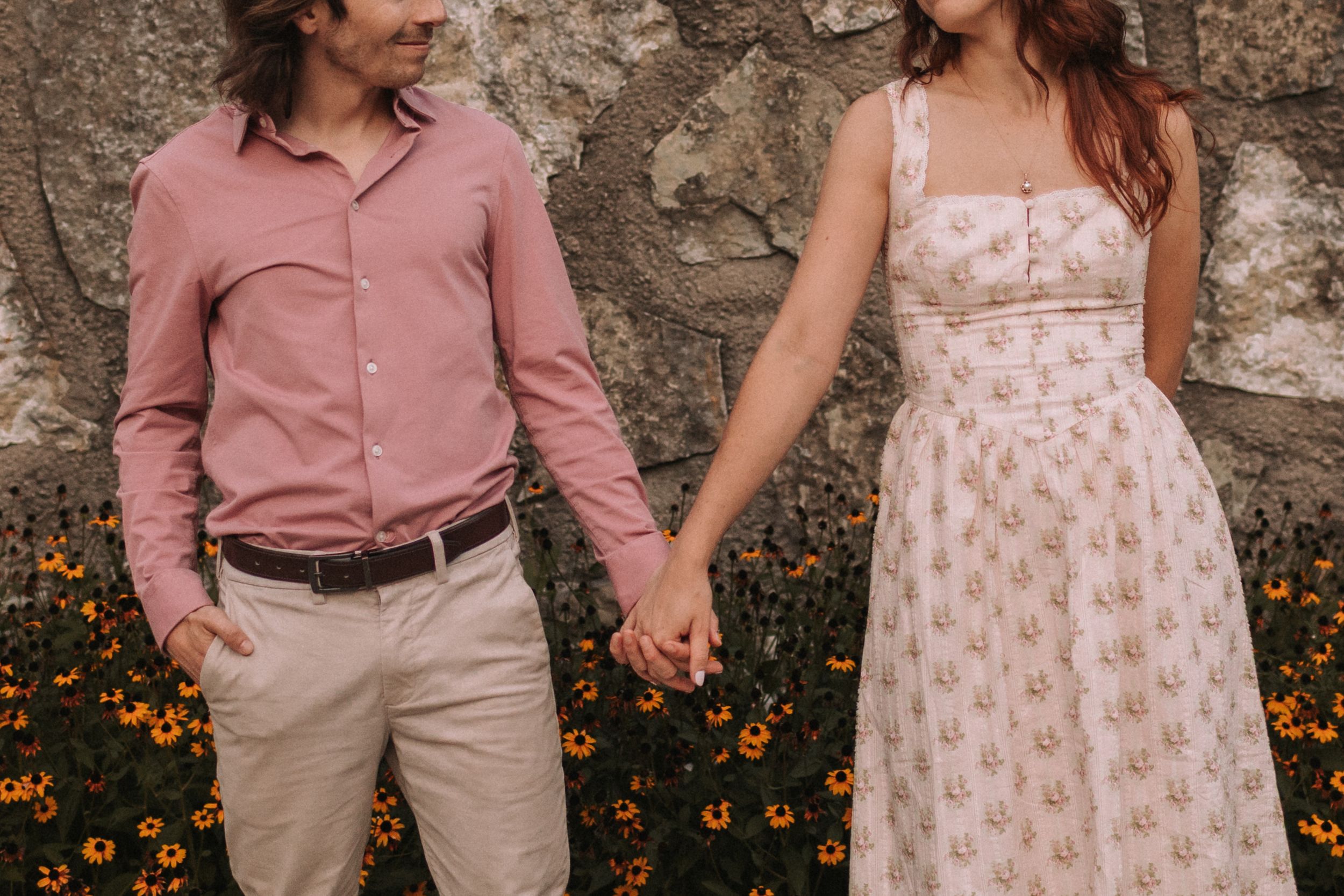 A couple holds hands against a stone wall with orange flowers blooming at the base.