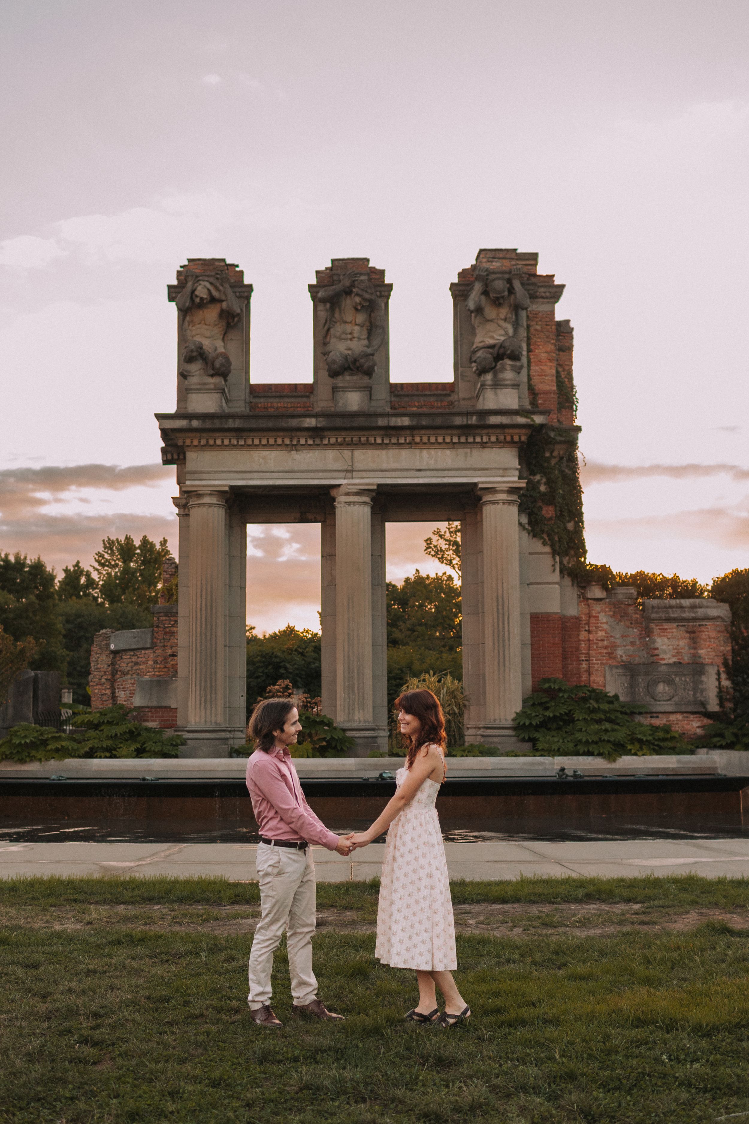 A romantic couple stands before a classical stone archway at sunset.