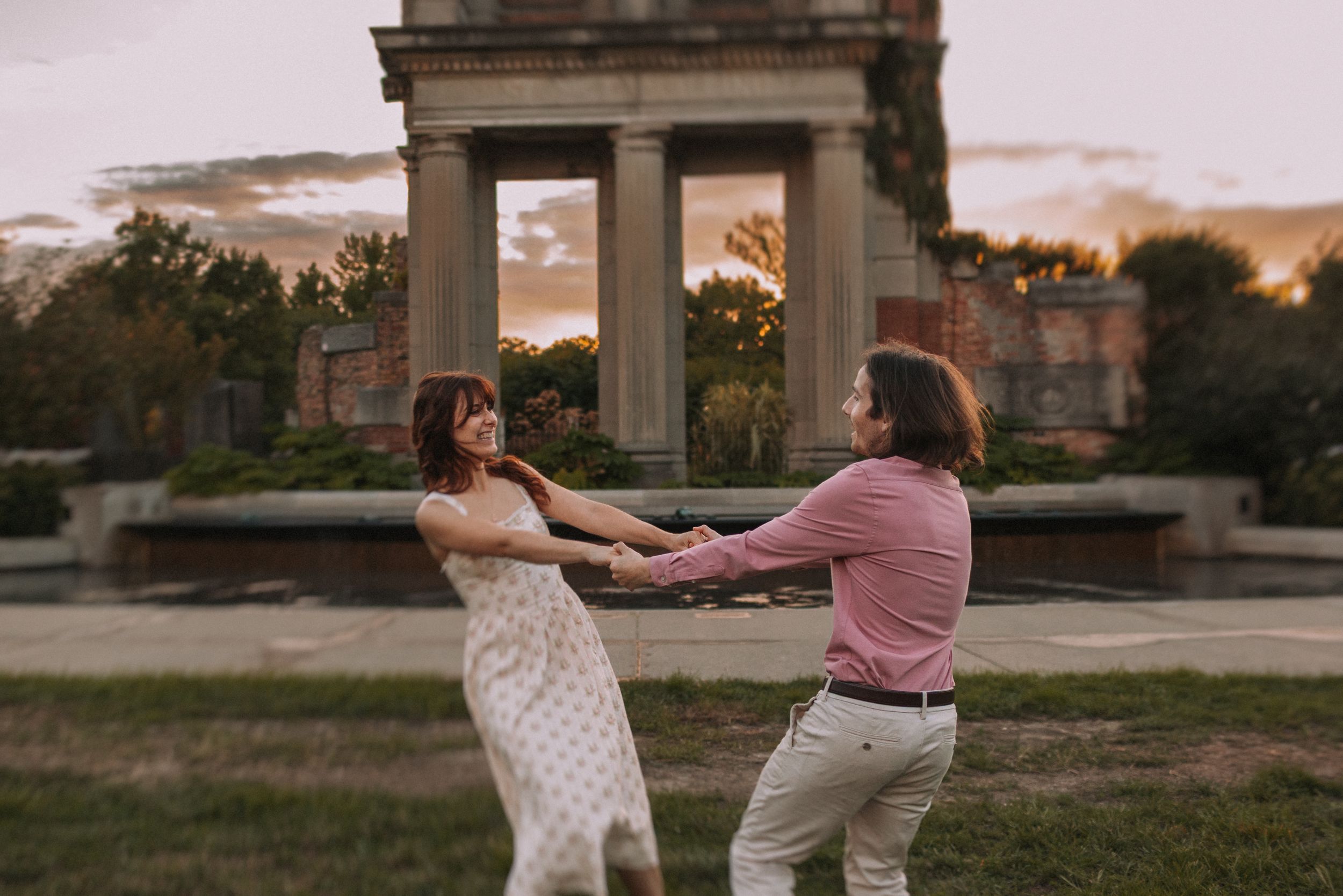 A couple dances playfully in front of ornate stone ruins at dusk.