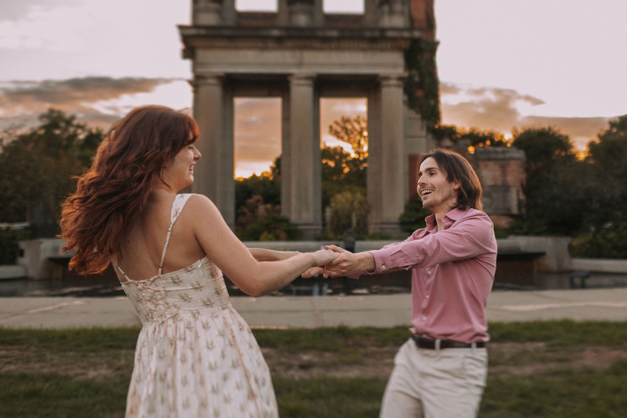 Two people dance together at sunset in a garden setting while wearing pink and white dresses.