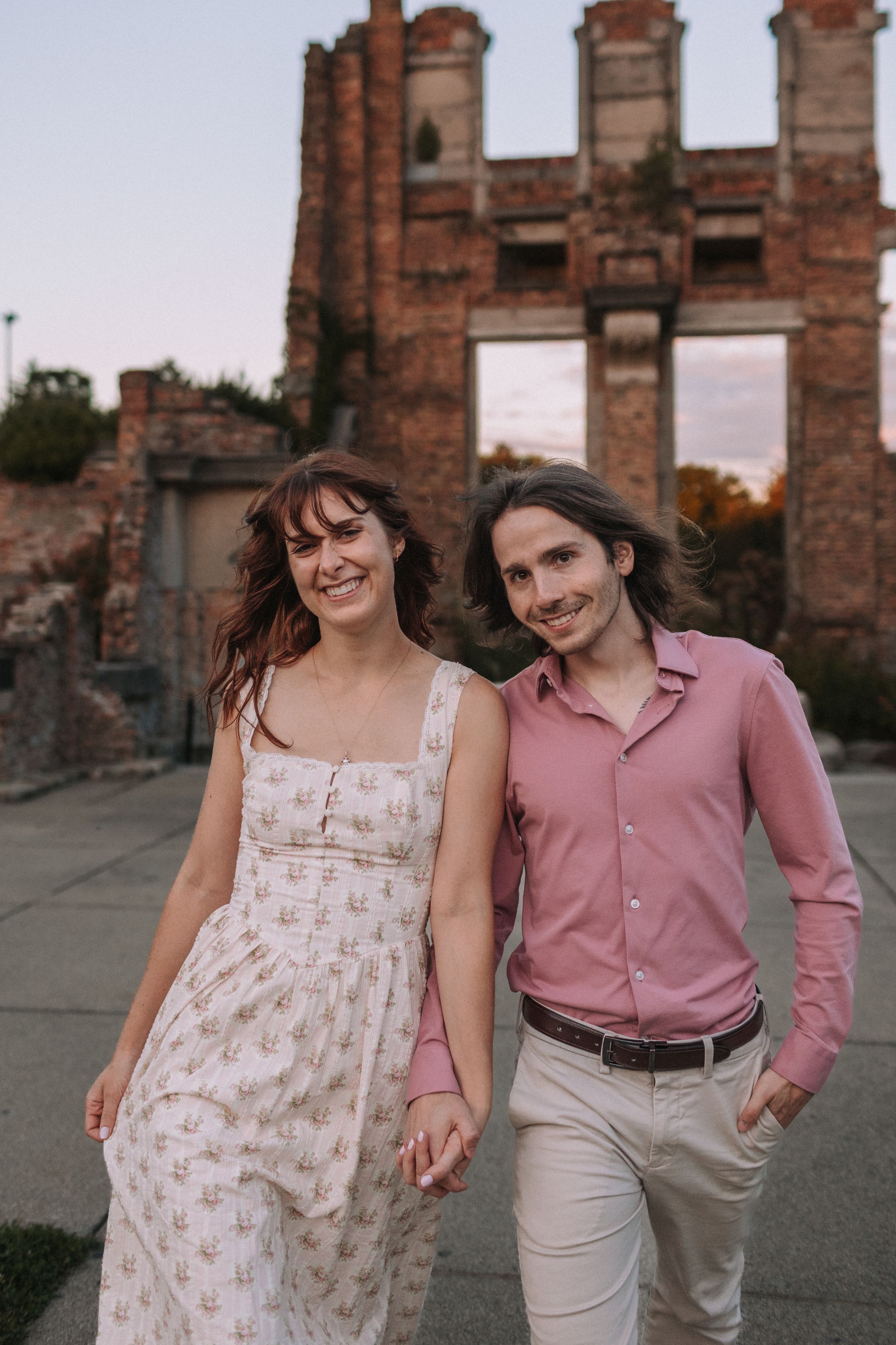 Romantic couple in pink and white outfits pose together against historic brick ruins at sunset.