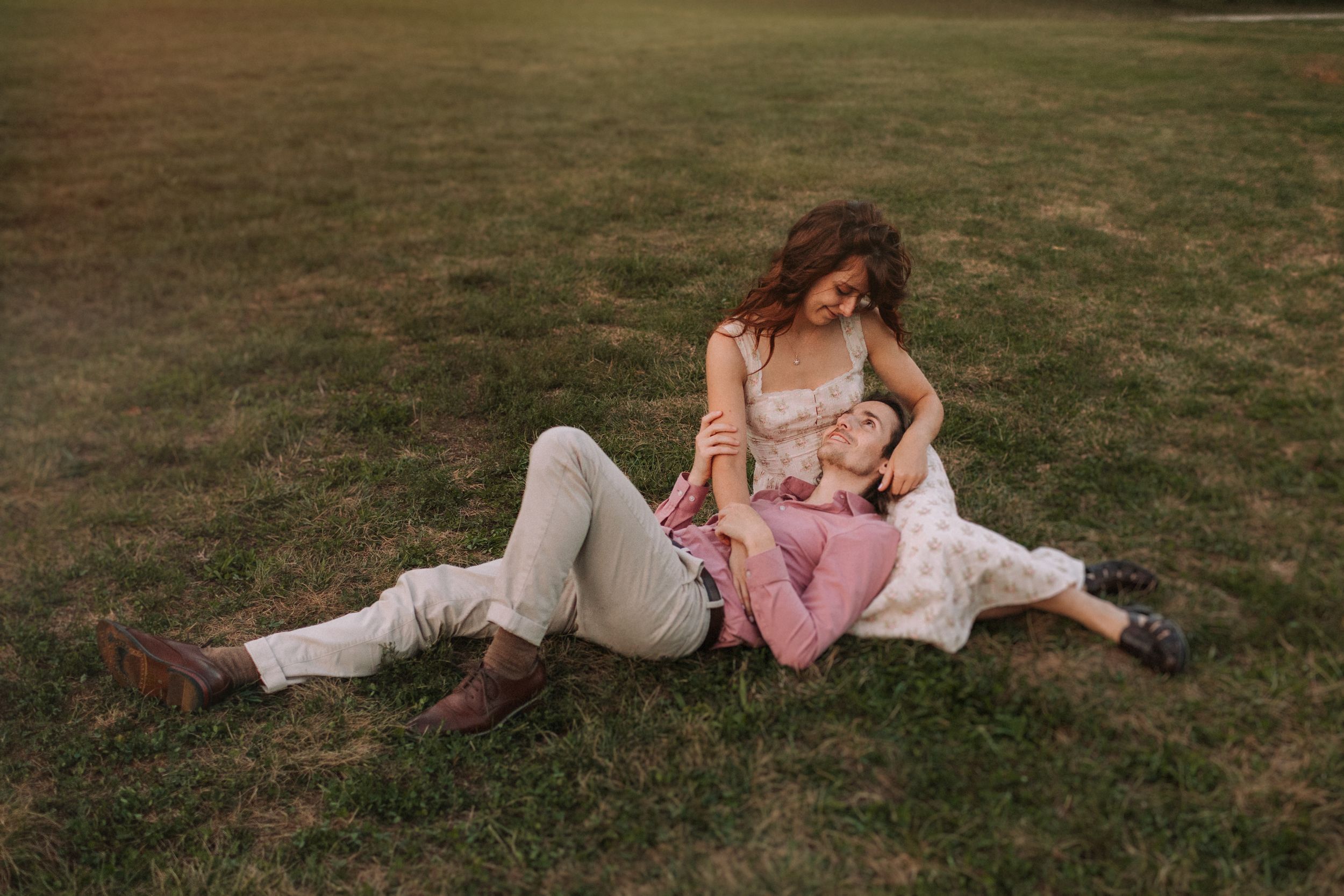 A couple lying together in a grassy field wearing romantic vintage-style clothing at sunset.