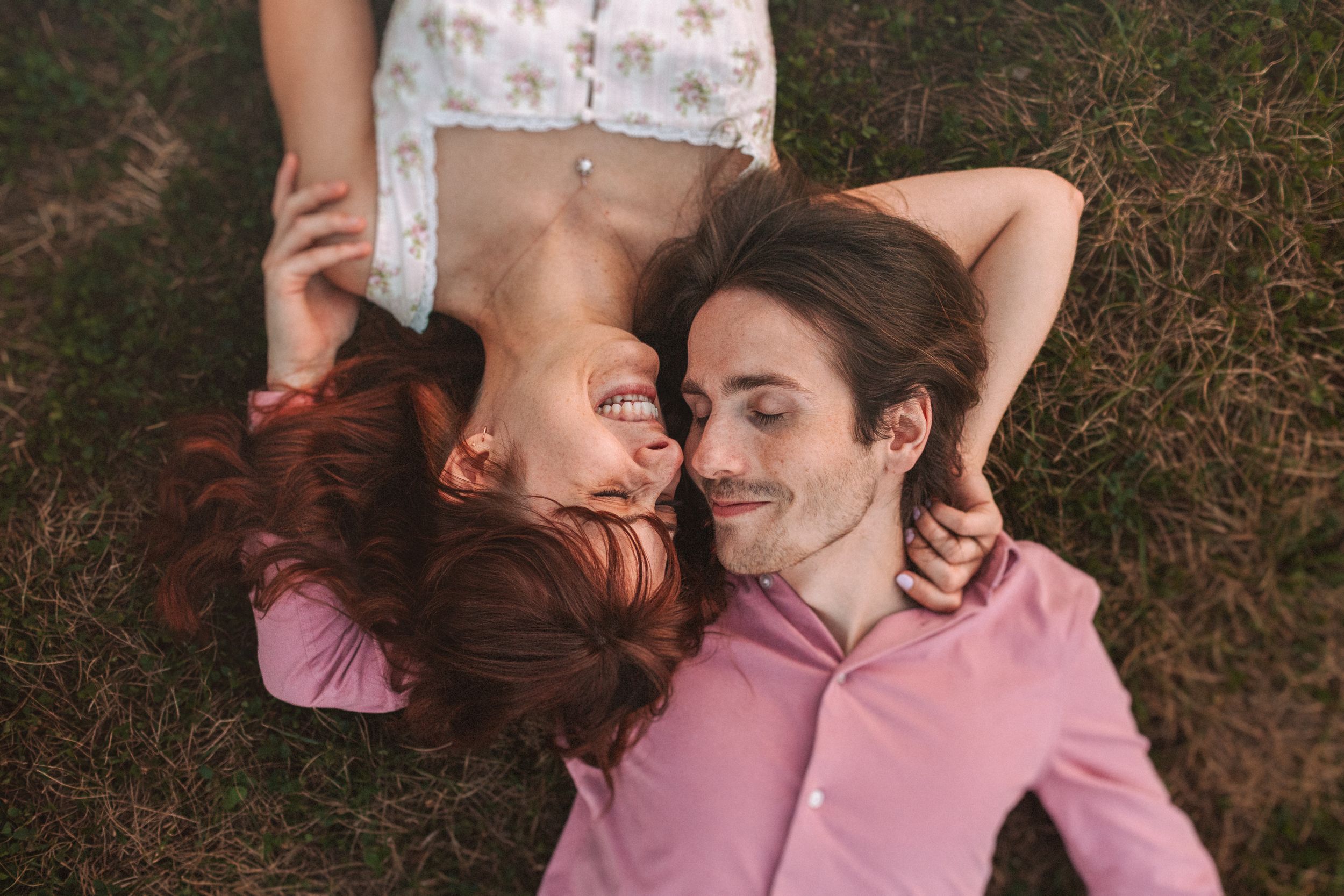 A couple in pink shirts lies on grass together while sharing a tender romantic moment outdoors.