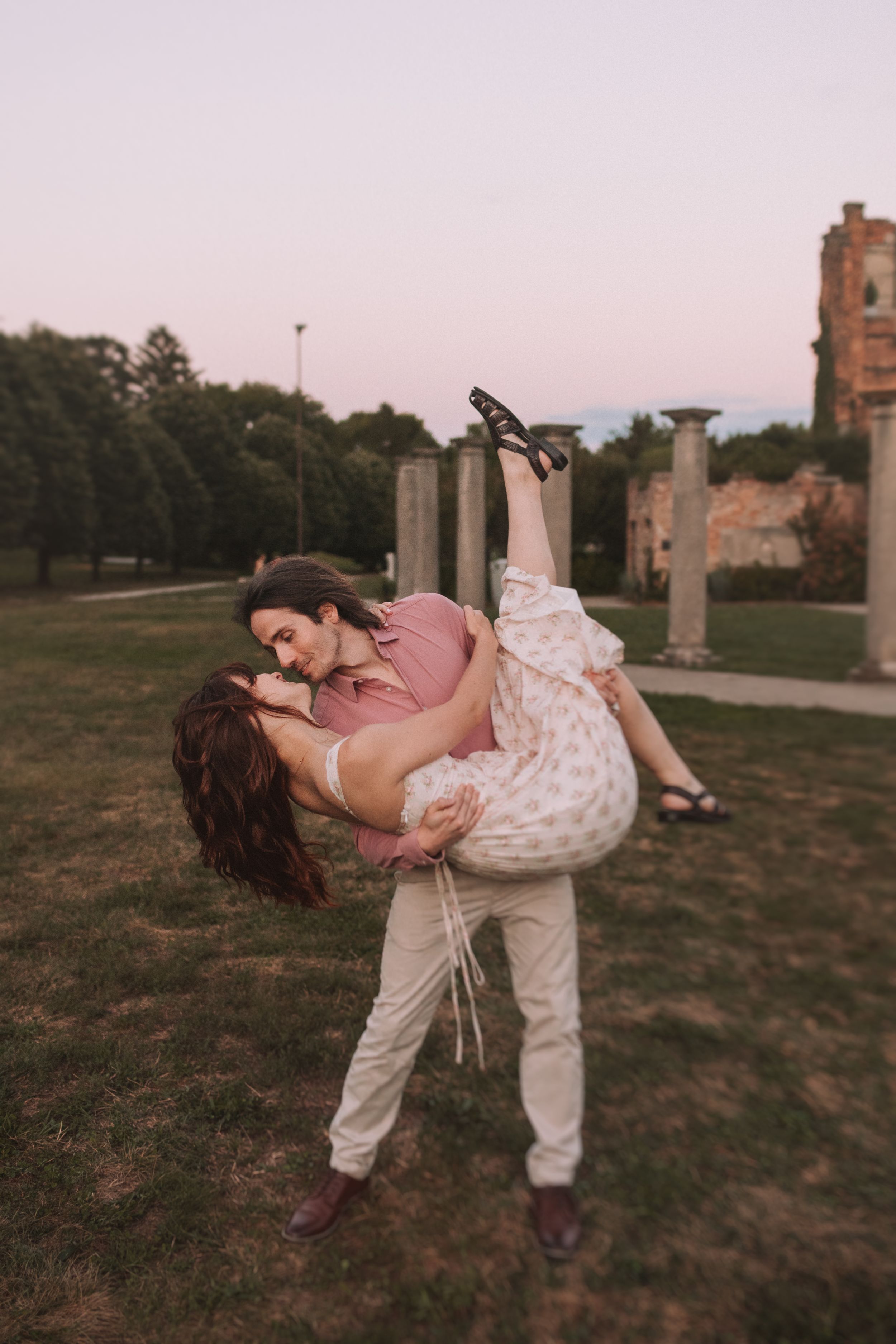 A couple in casual attire dances playfully in a field at sunset with stone columns in the background.