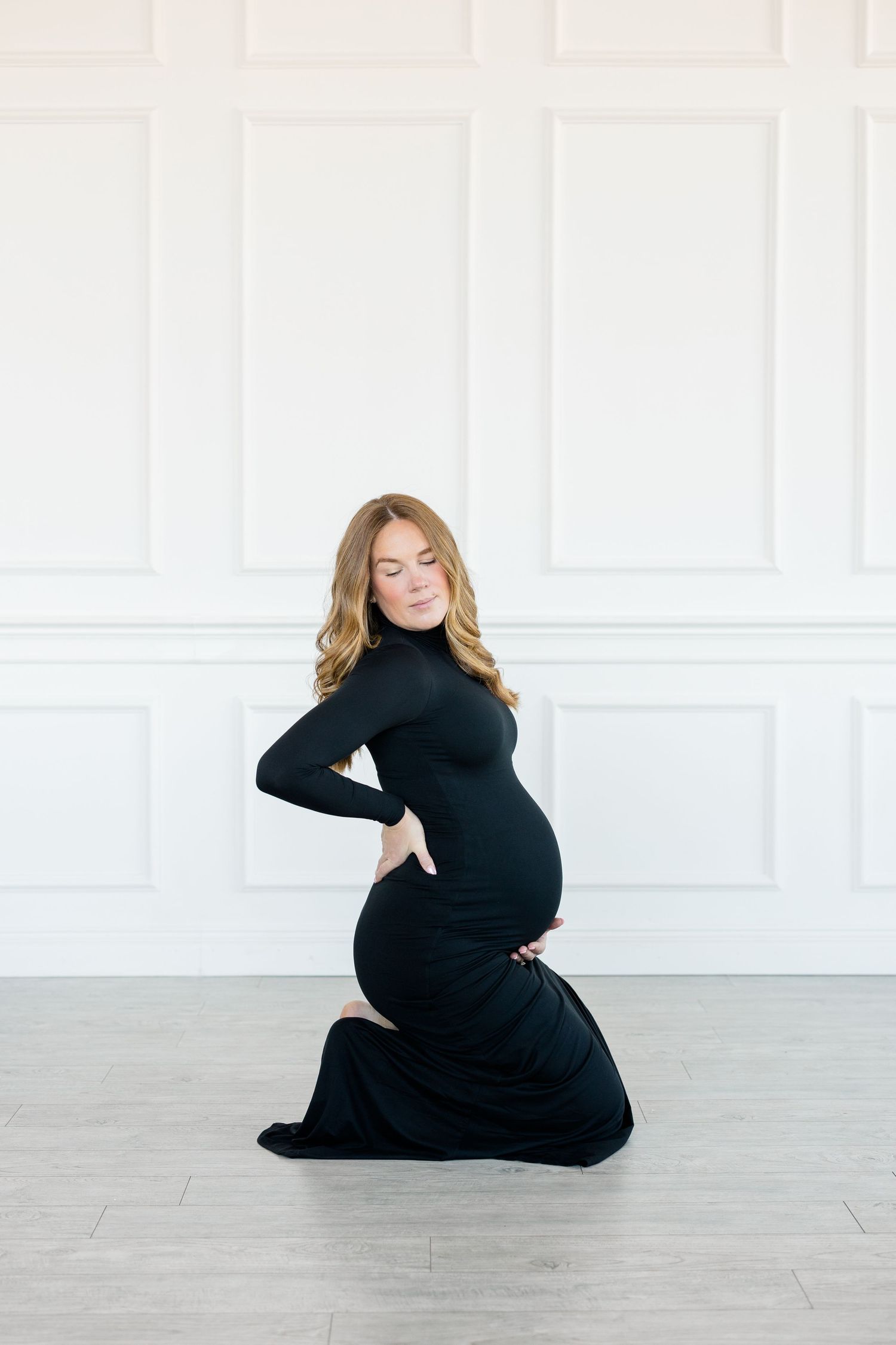 A pregnant woman in a black turtleneck dress poses gracefully on a light gray floor in a minimalist white room.