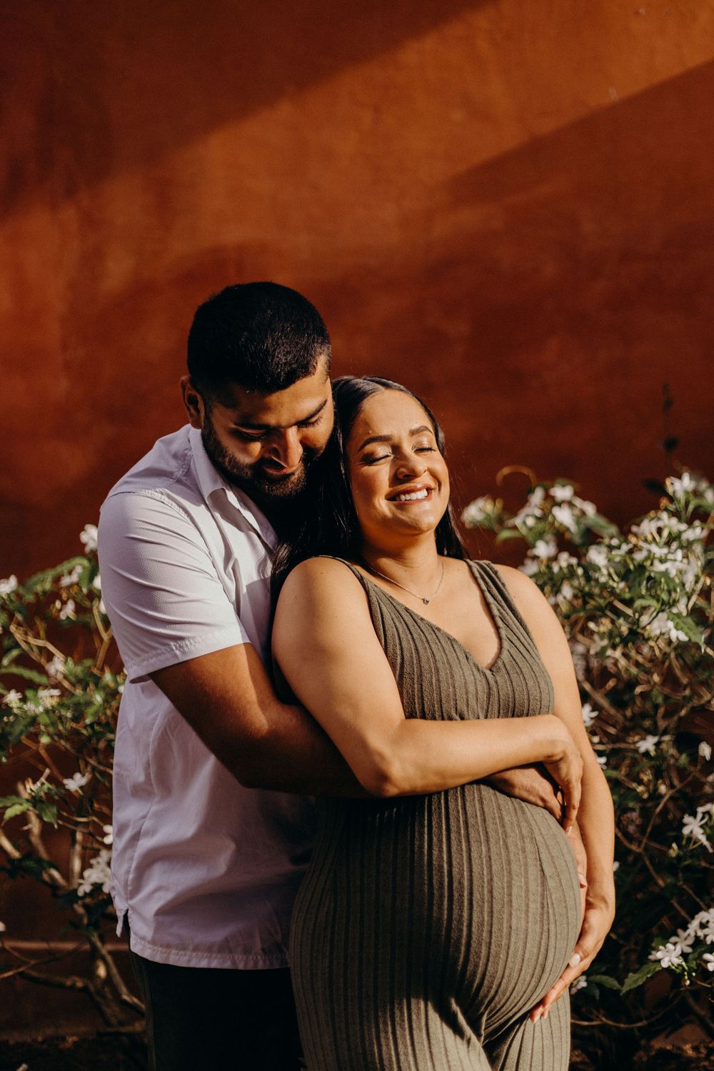 Maternity photoshoot featuring a couple embracing in front of flowering bushes at sunset.