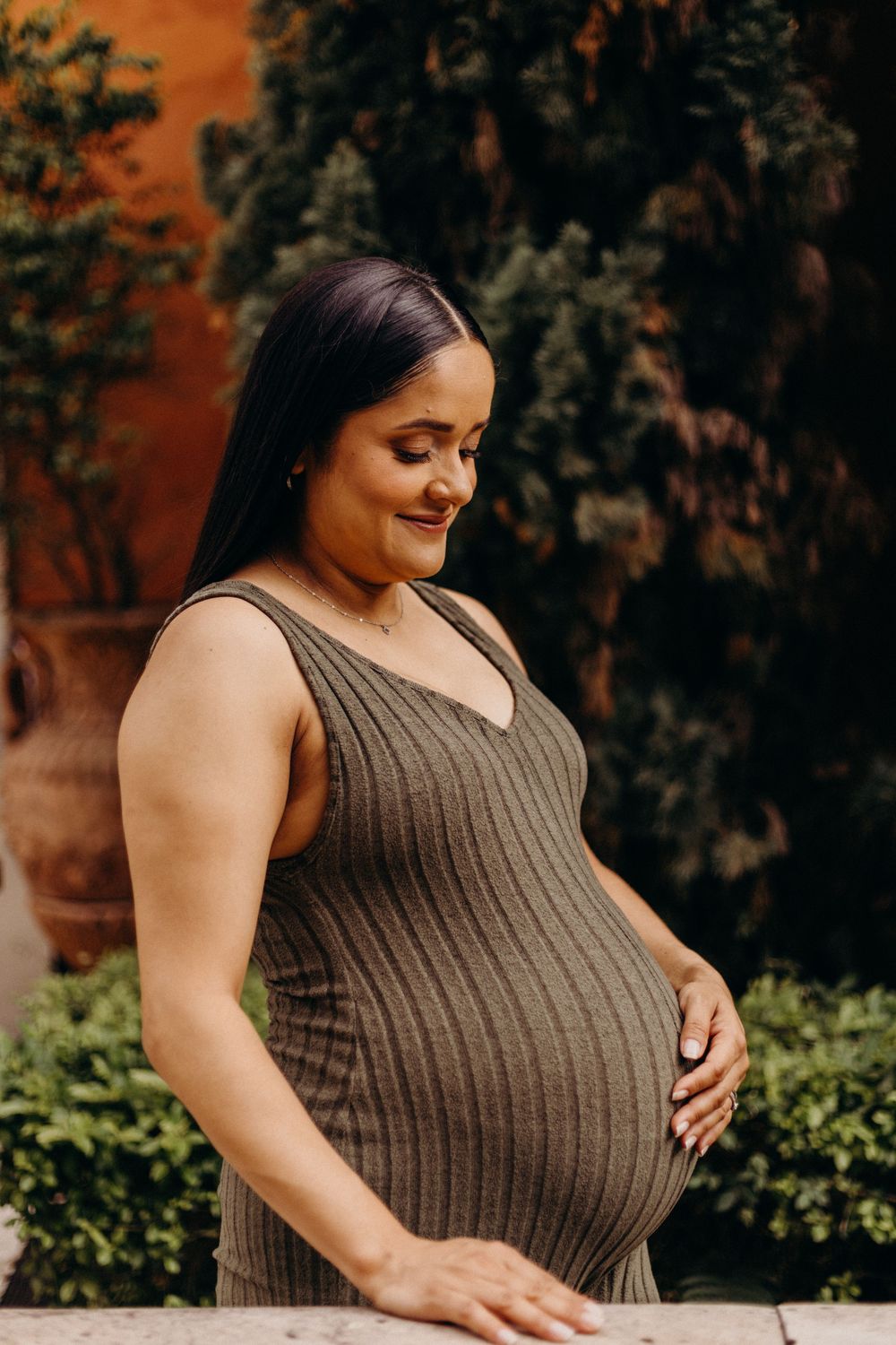 Expectant mother in brown ribbed dress poses for maternity photos in garden setting.
