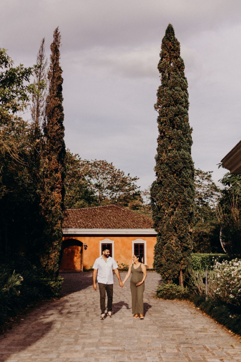 Couple walking together on stone pathway with tall cypress trees and orange building in background.