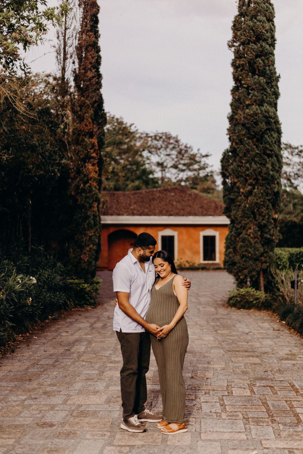 Couple walks hand in hand along a stone pathway lined with cypress trees and orange building.