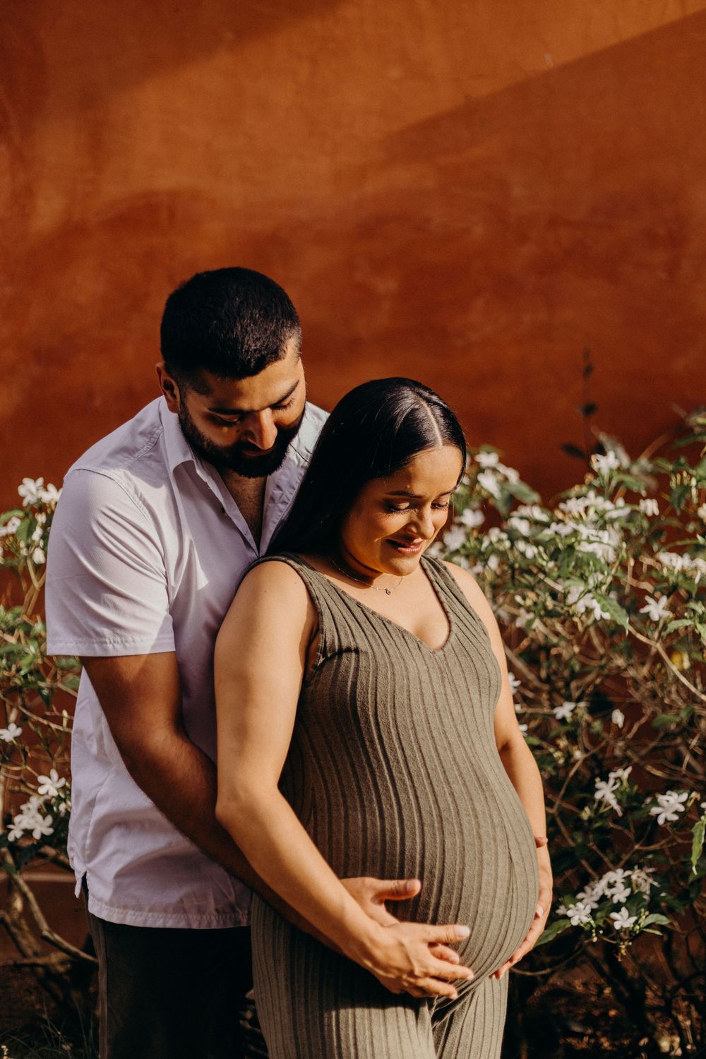 Intimate maternity photo session showing a couple embracing near white flowers against a terracotta wall.