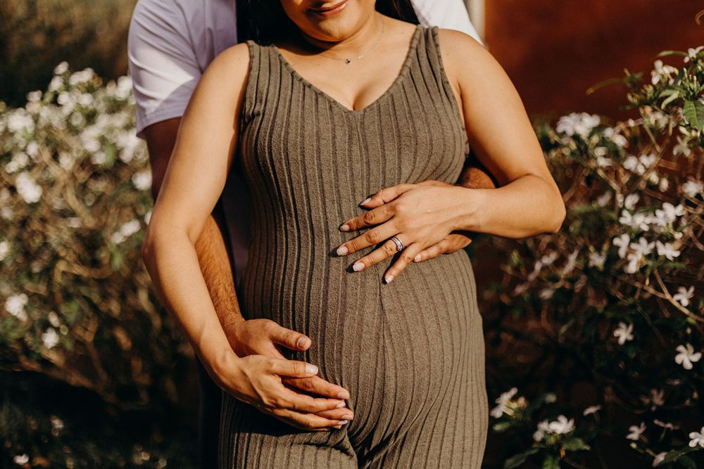 Maternity photo series showing pregnant belly in brown ribbed dress against sunlit garden with white flowers.