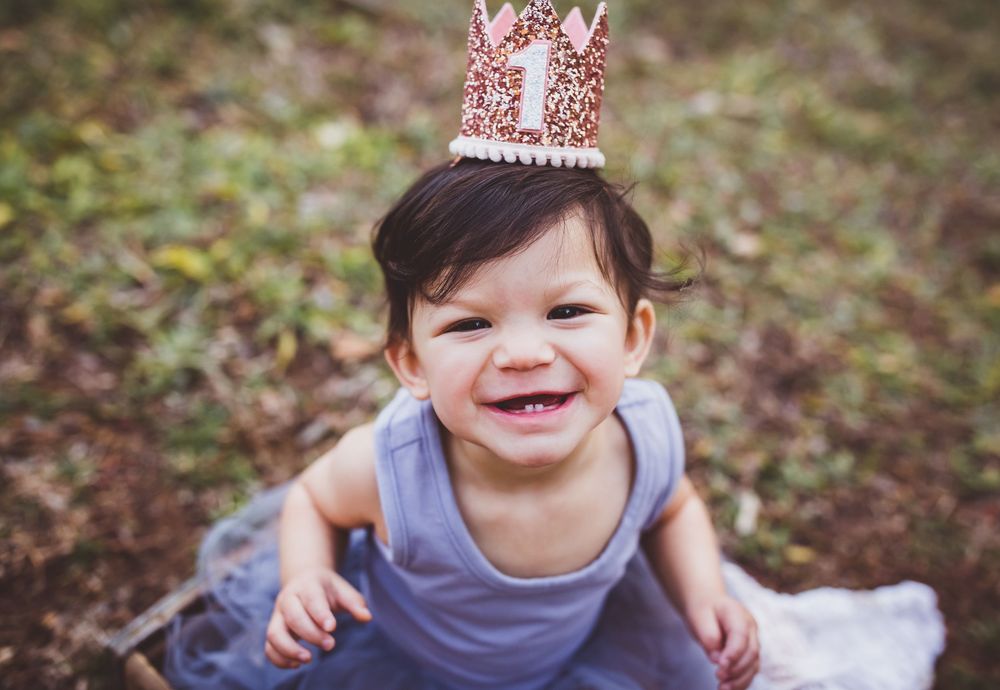 A baby wearing a pink crown sits on a white blanket in an outdoor setting.