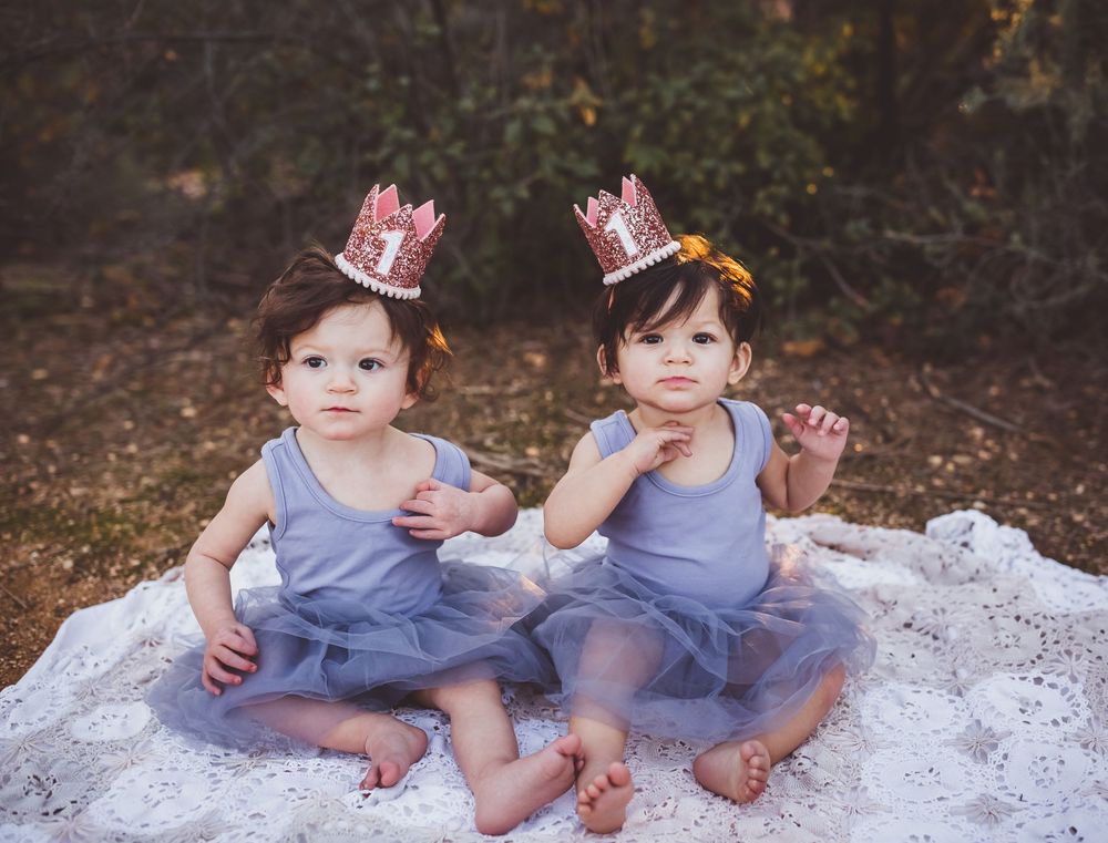 Two children in lavender dresses and pink crowns sit together on white fabric during an outdoor photo session.