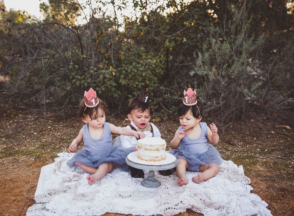 Children gather around a birthday cake on a white lace blanket in a natural outdoor setting.
