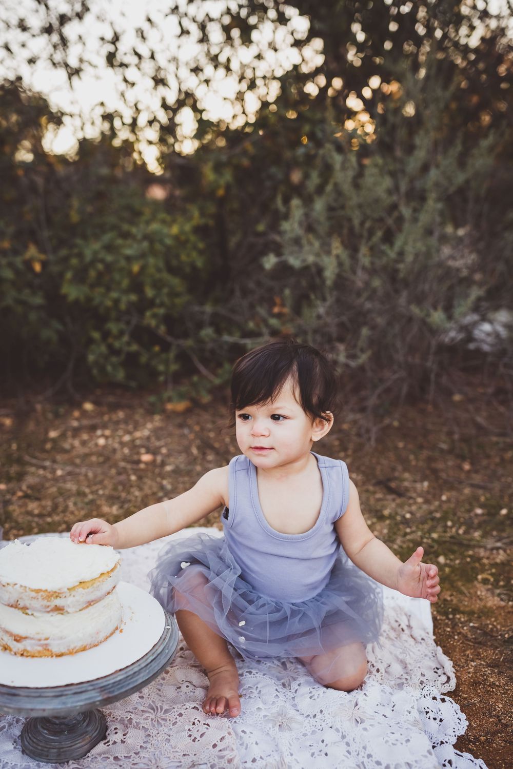 A small child in a blue dress sits on lace fabric near a white cake during a golden hour photoshoot.