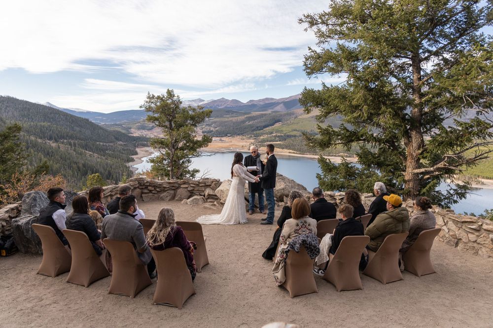 Outdoor wedding ceremony guests seated on brown chairs overlooking a scenic mountain lake landscape.