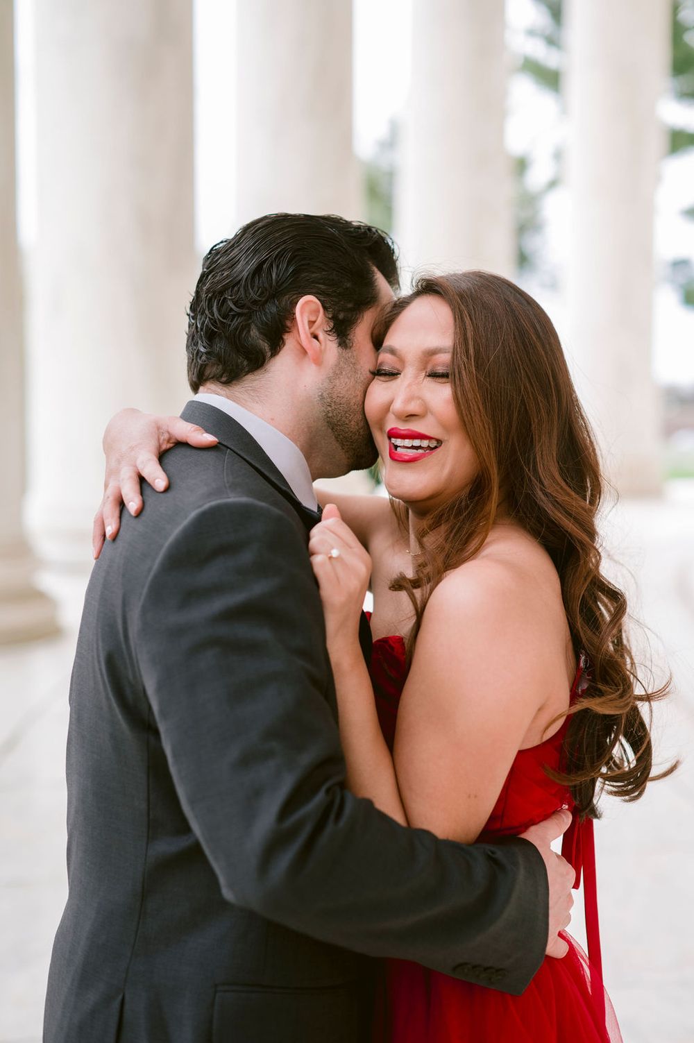 A romantic couple embraces and shares a tender moment between elegant white columns.