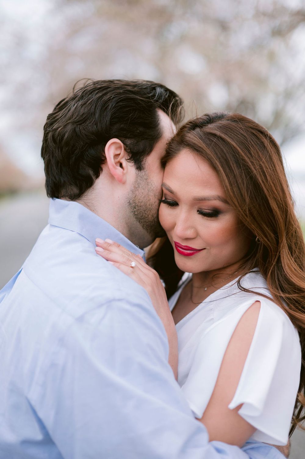 A romantic couple embraces outdoors in soft lighting, with the woman wearing white and the man in a light blue shirt.