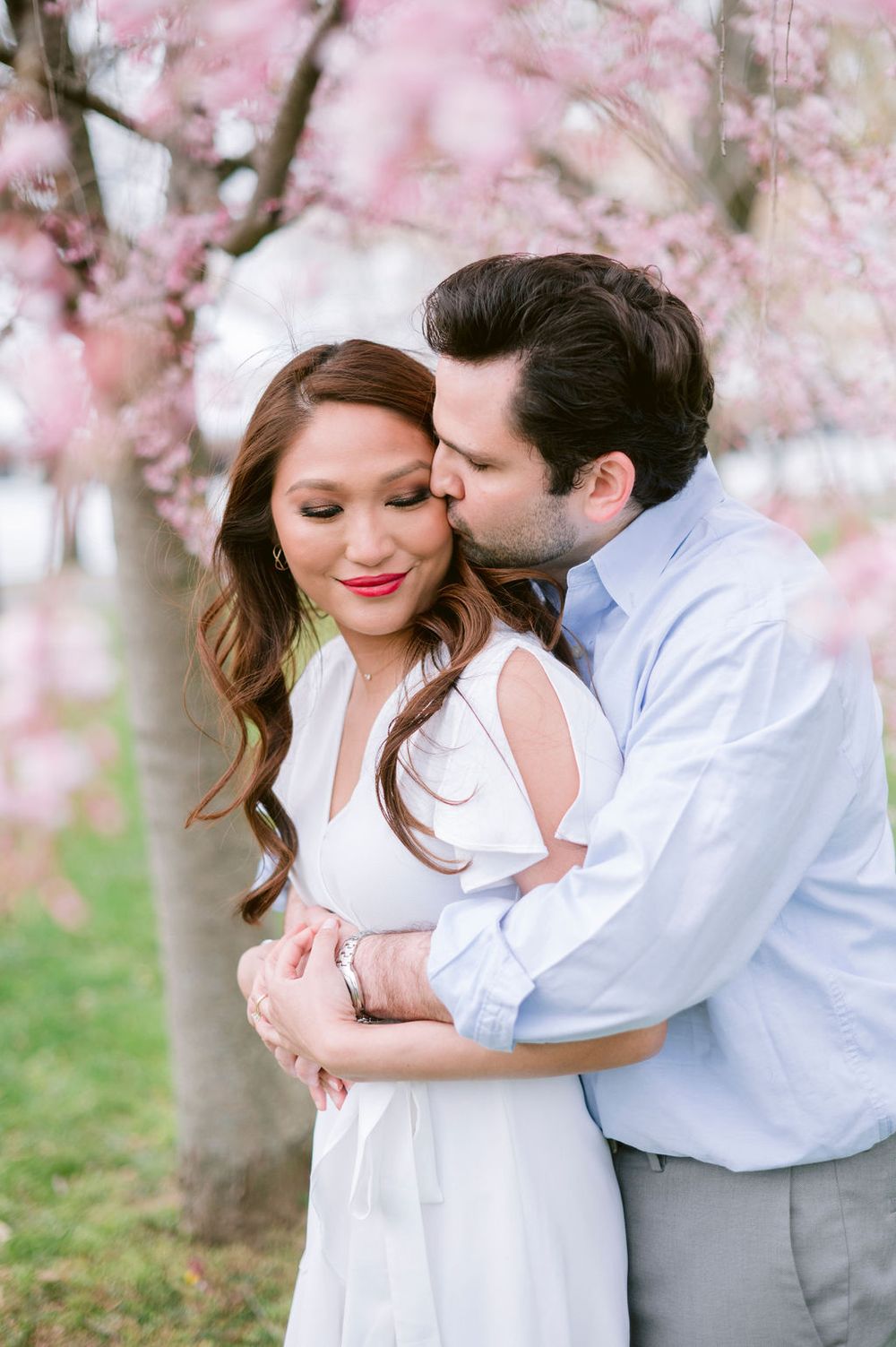 Romantic couple embraces under blooming cherry blossom trees during spring engagement photo session.
