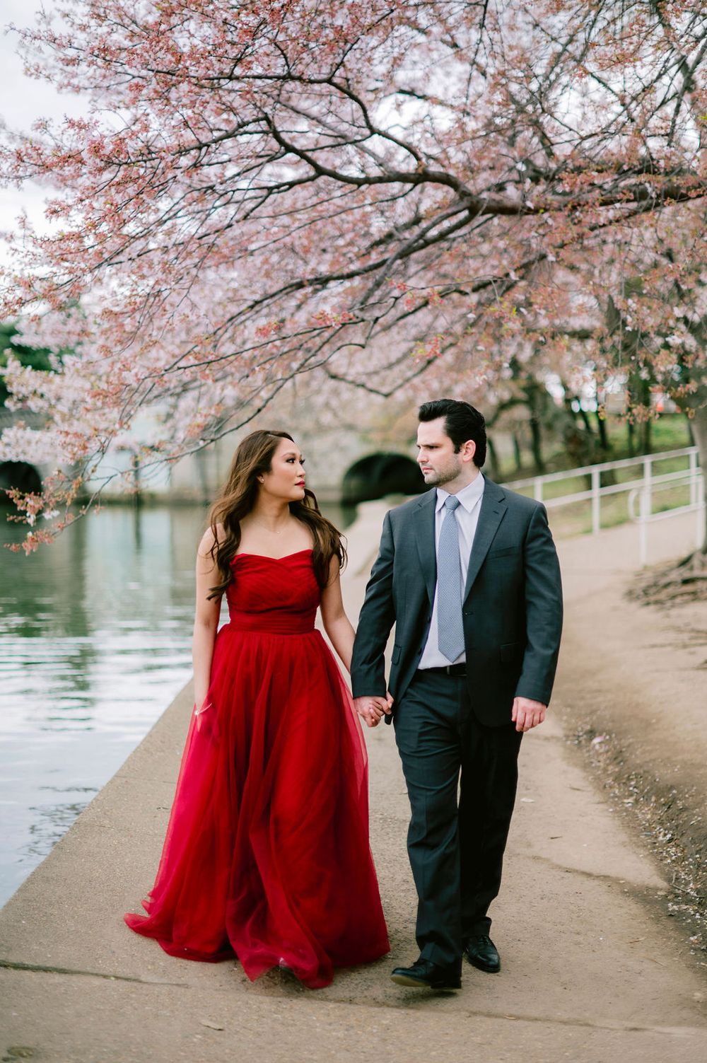 A couple in formal attire walks beneath blooming cherry blossoms along a waterfront path.