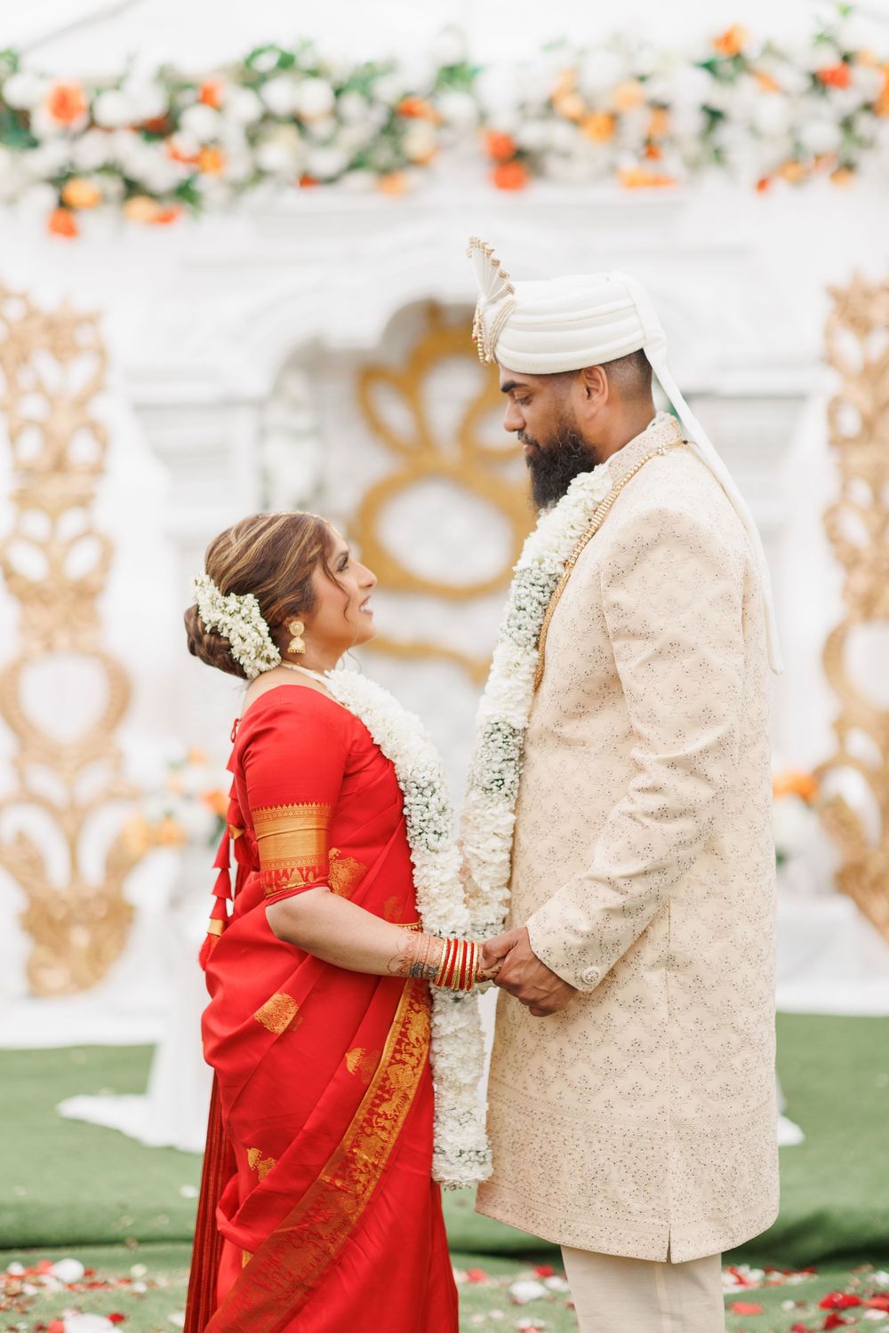 A couple in traditional wedding attire, with the bride in a red sari and groom in cream sherwani, share joyful moments together.