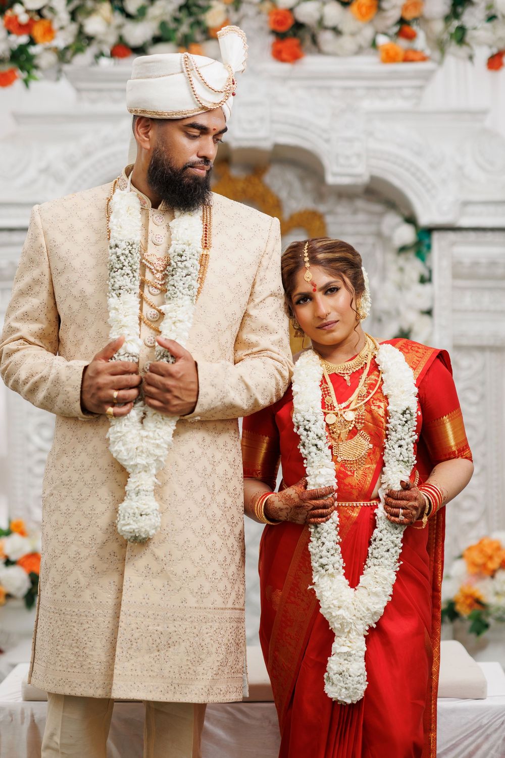 Traditional Indian wedding ceremony showing couple exchanging garlands in red and cream outfits with floral backdrop.