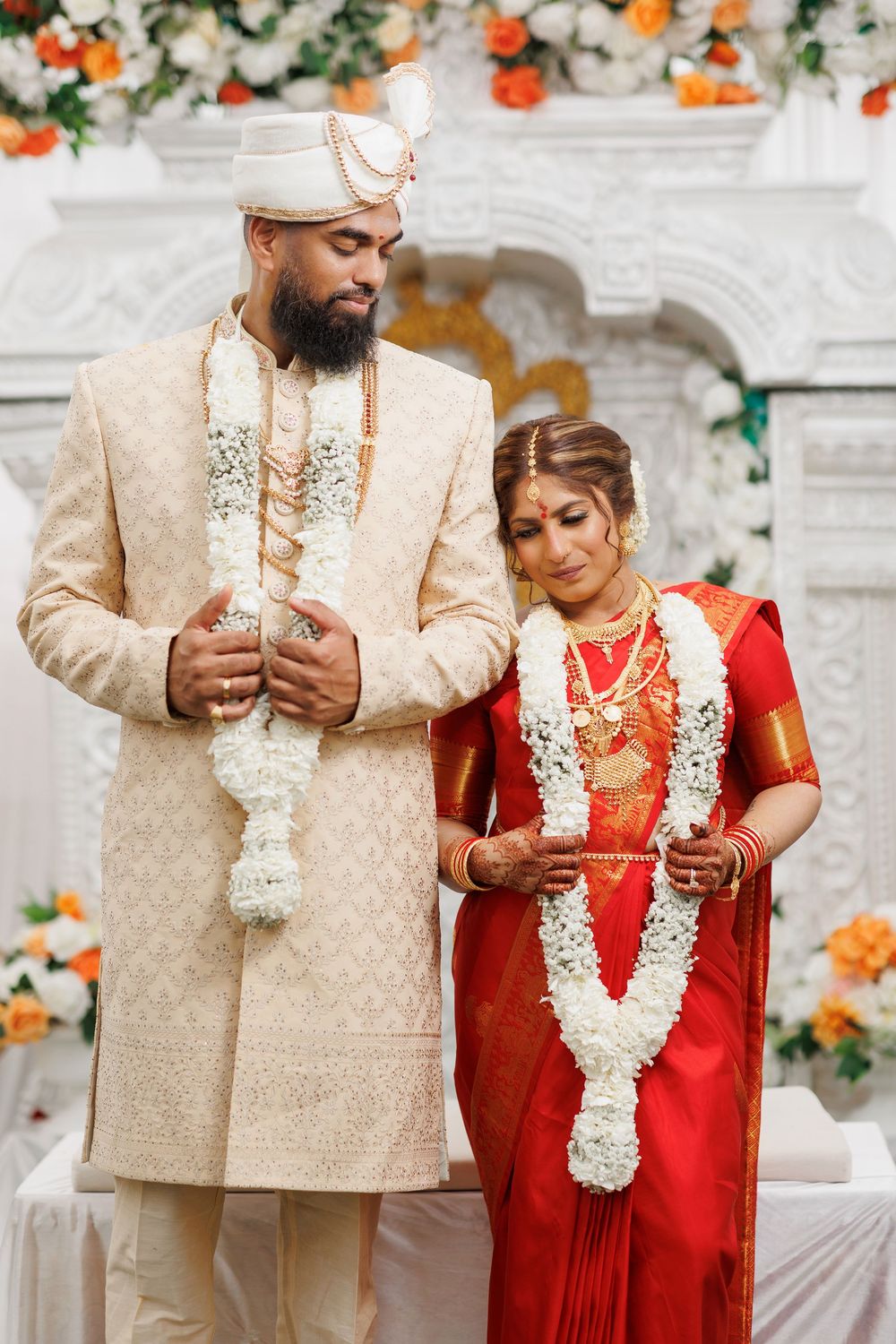 A couple in traditional South Asian wedding attire exchange garlands during a ceremony decorated with orange flowers.