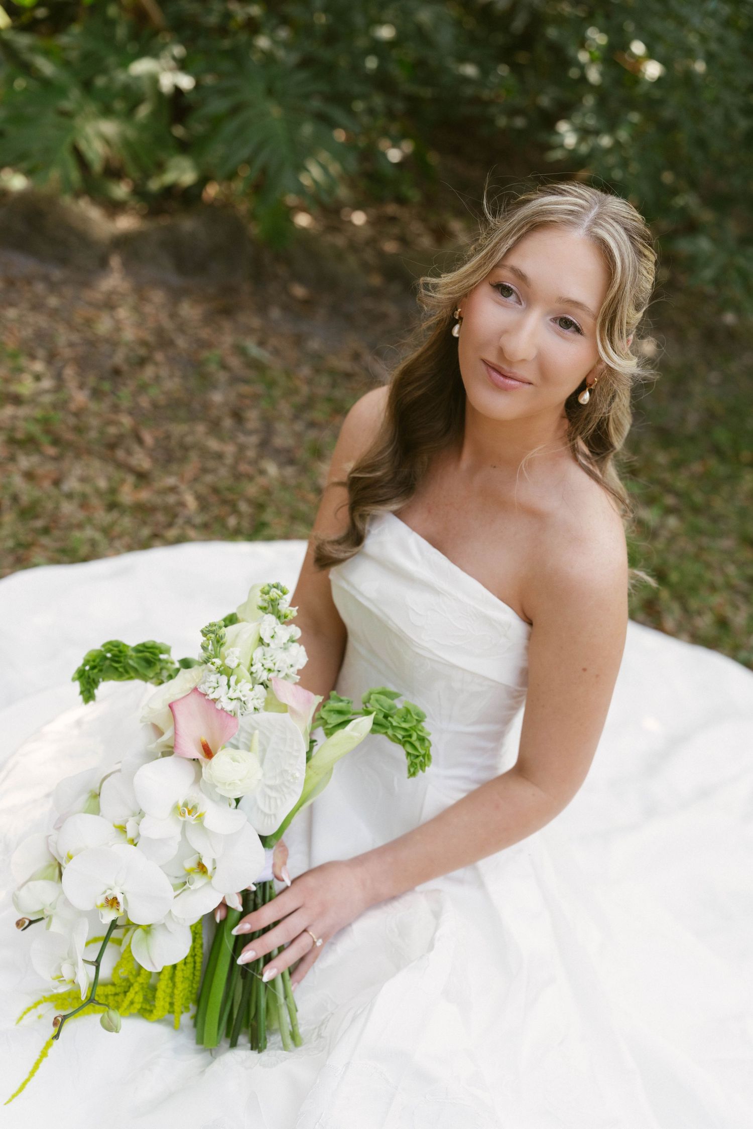 Smiling bride in strapless white gown holding elegant bouquet of white orchids and pink flowers outdoors.