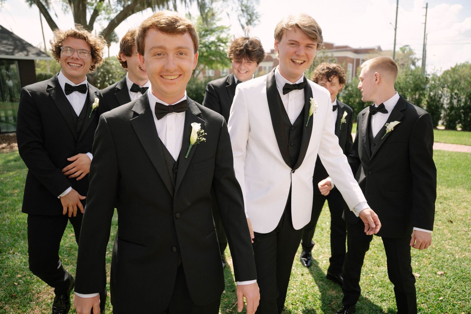 Groom in white tuxedo jacket stands with groomsmen in black tuxedos outdoors on green lawn.