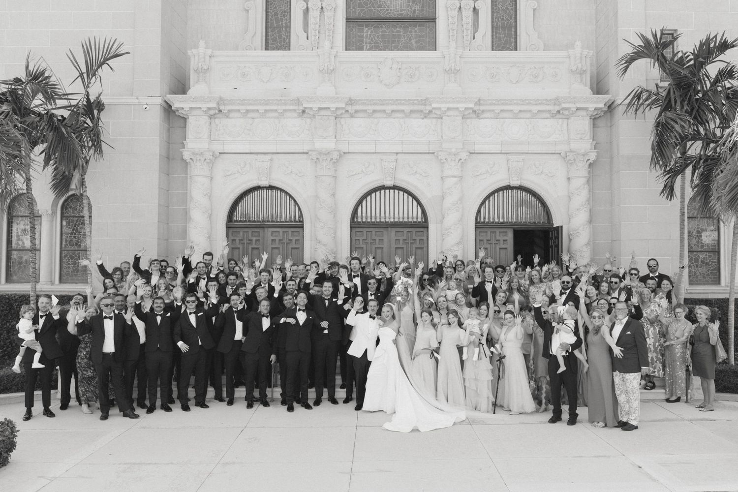 Black and white wedding photo of bride, groom, and large guest group posed outside ornate building with arches.