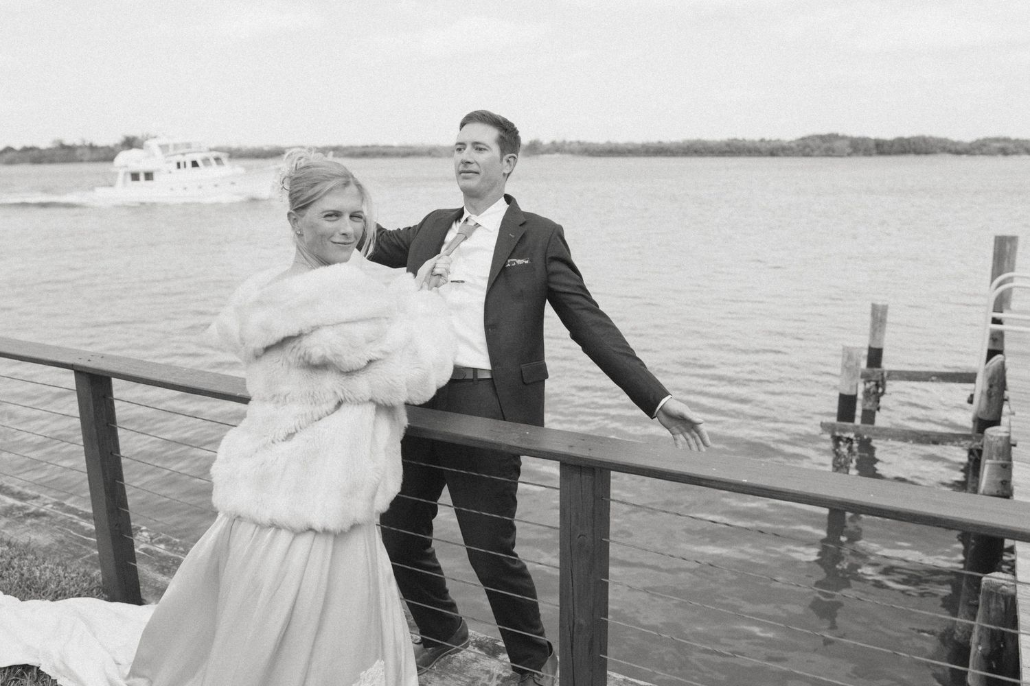 Black and white photo of a bride and groom posing on a waterfront dock, with a boat visible on the water behind them.