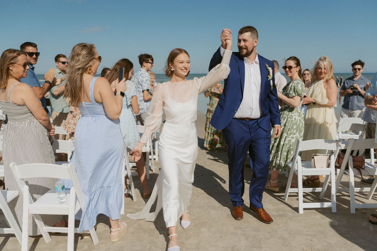 Joyful newlyweds walk down beach aisle hand-in-hand, bride in white gown, groom in blue suit, guests cheering.