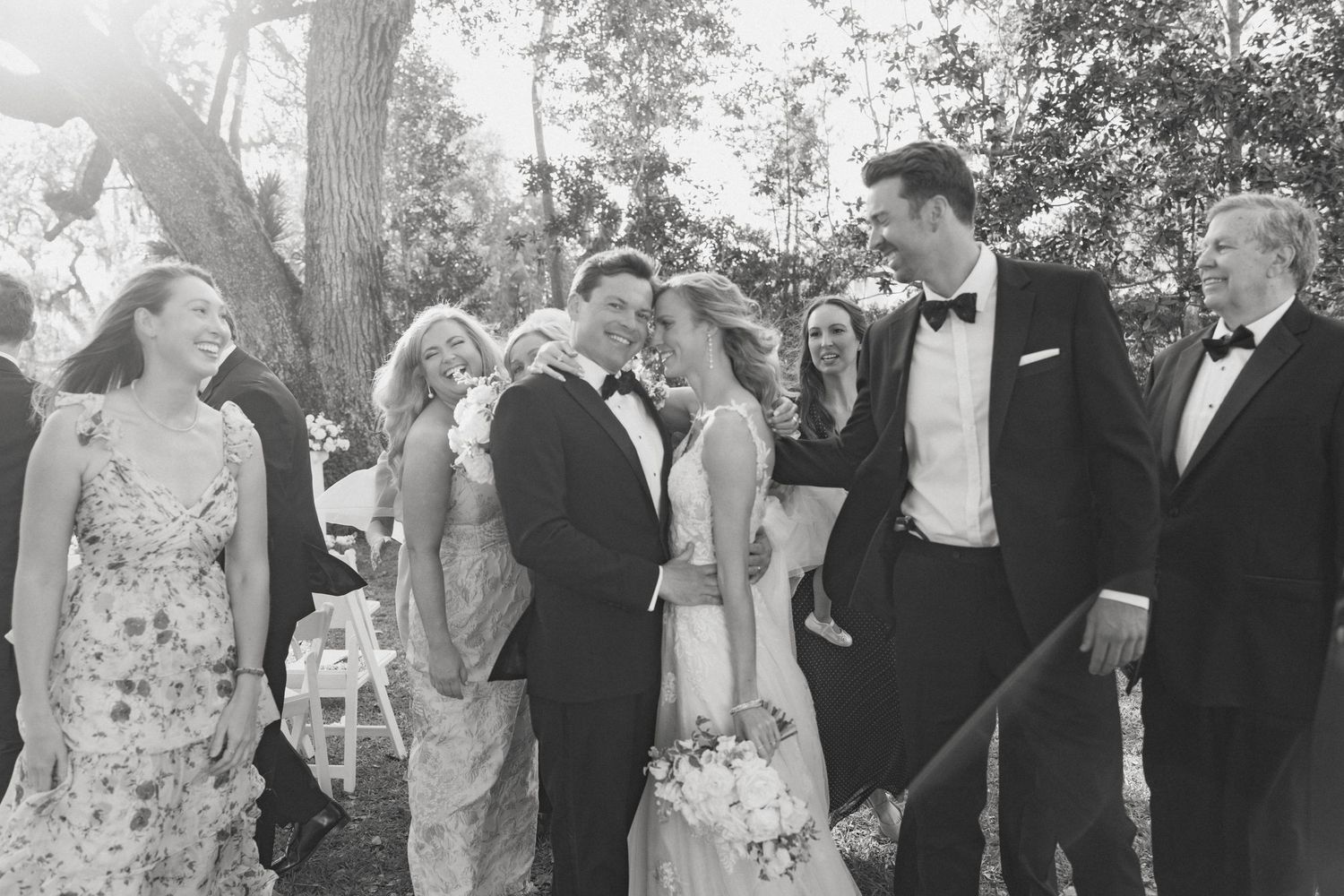 Black and white wedding photo of bride and groom surrounded by smiling guests outdoors under trees.
