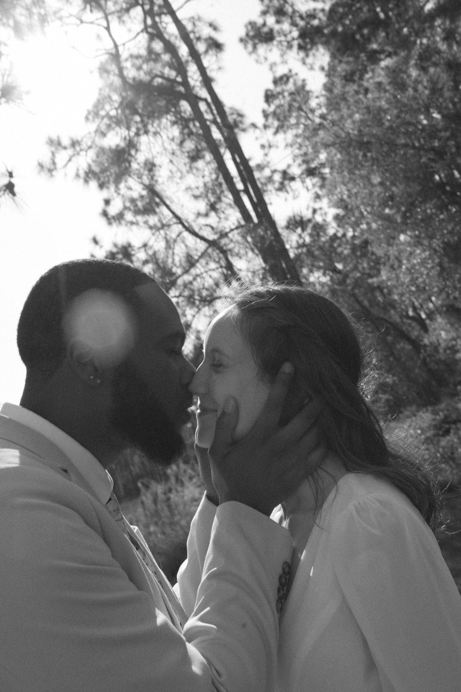 Black and white photo of a couple sharing a tender moment outdoors, man gently holding womans face near trees.