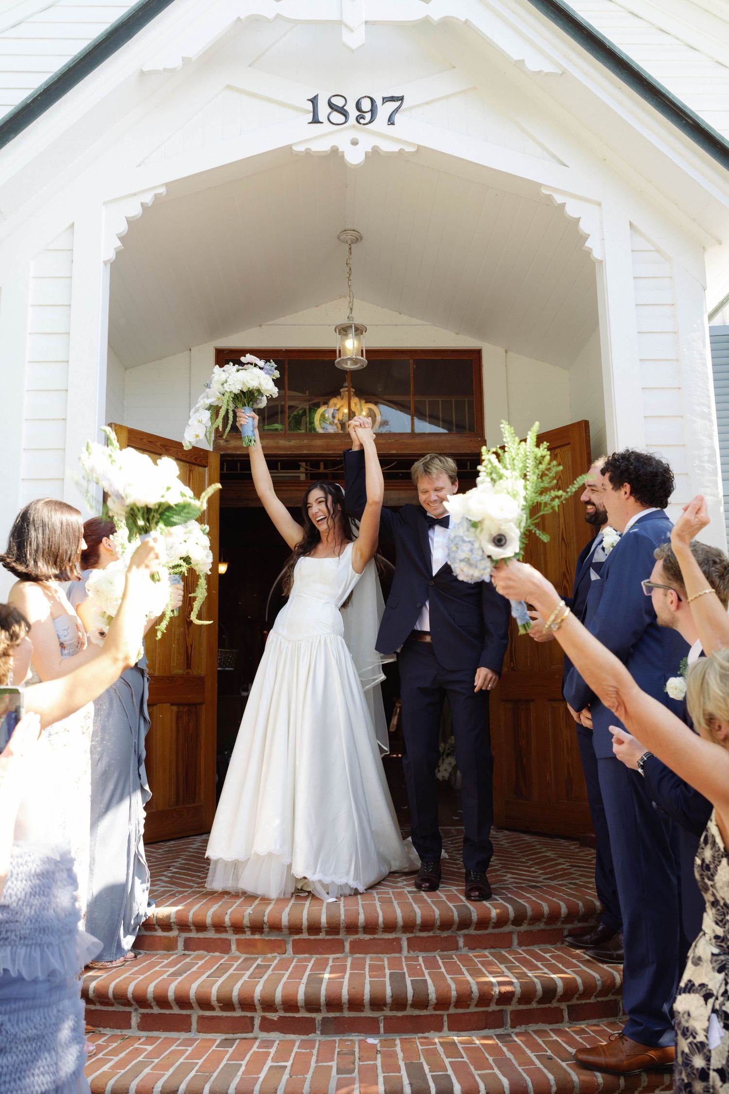 Bride and groom exit a historic 1897 white chapel, raising bouquets as guests cheer on the brick steps.