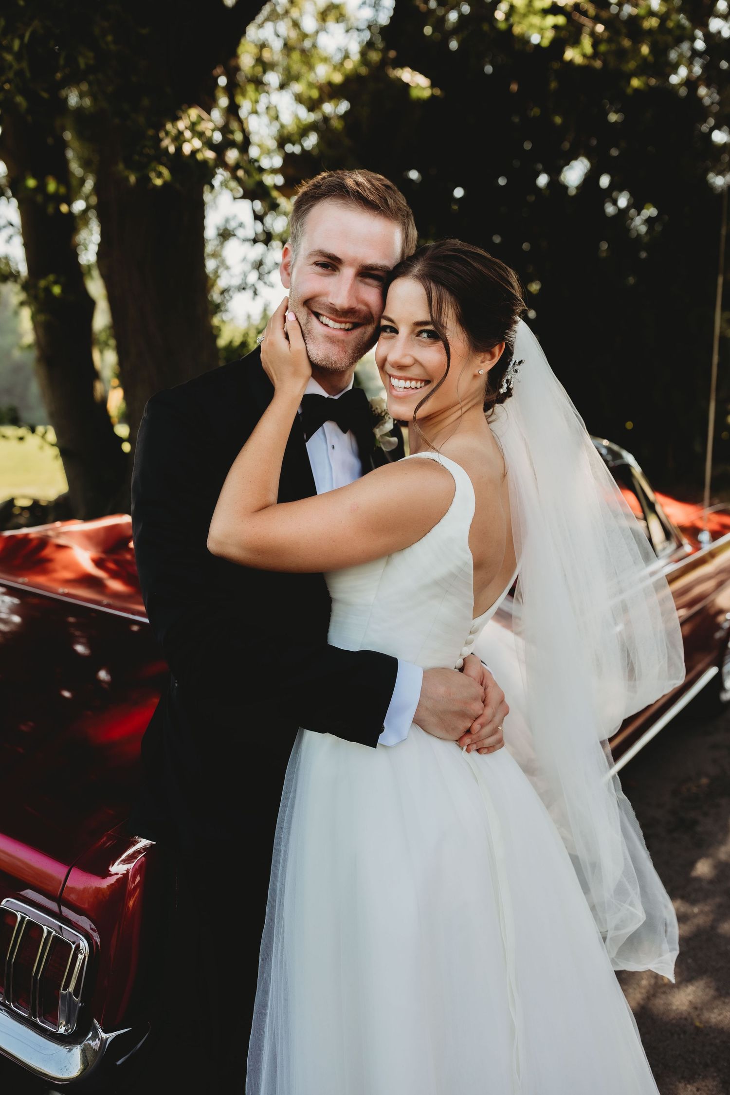 A joyful couple shares intimate moments on their wedding day near a vintage red car in a sun-dappled outdoor setting.