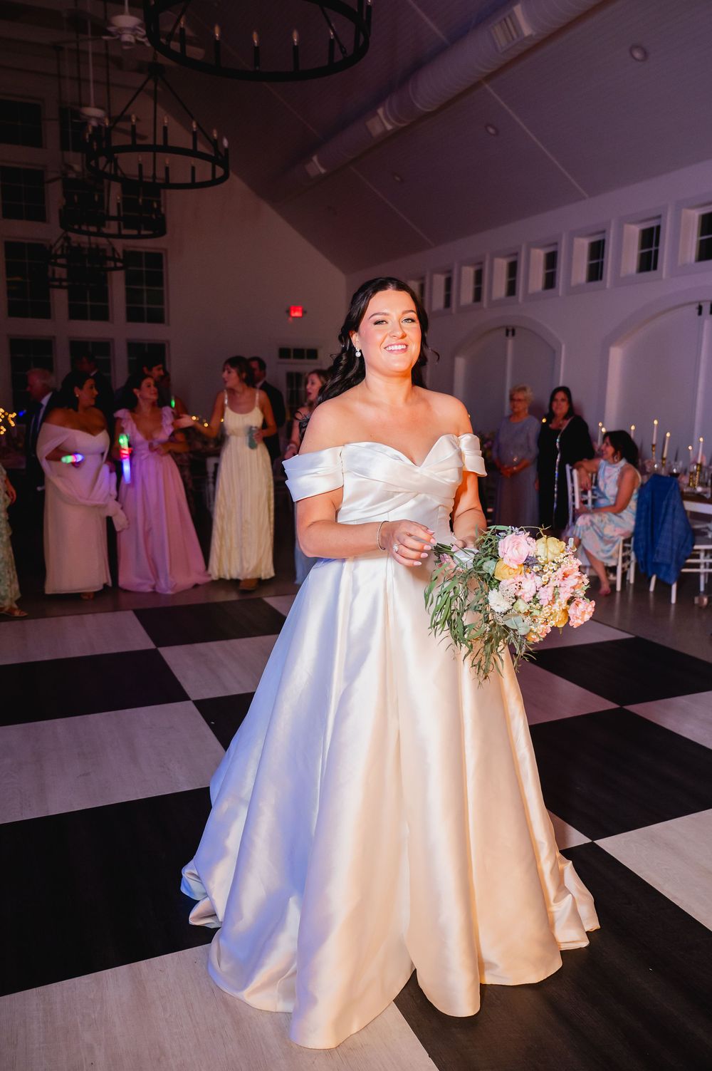 A bride in a stunning off-shoulder satin gown holds her bouquet on a black and white checkered floor.