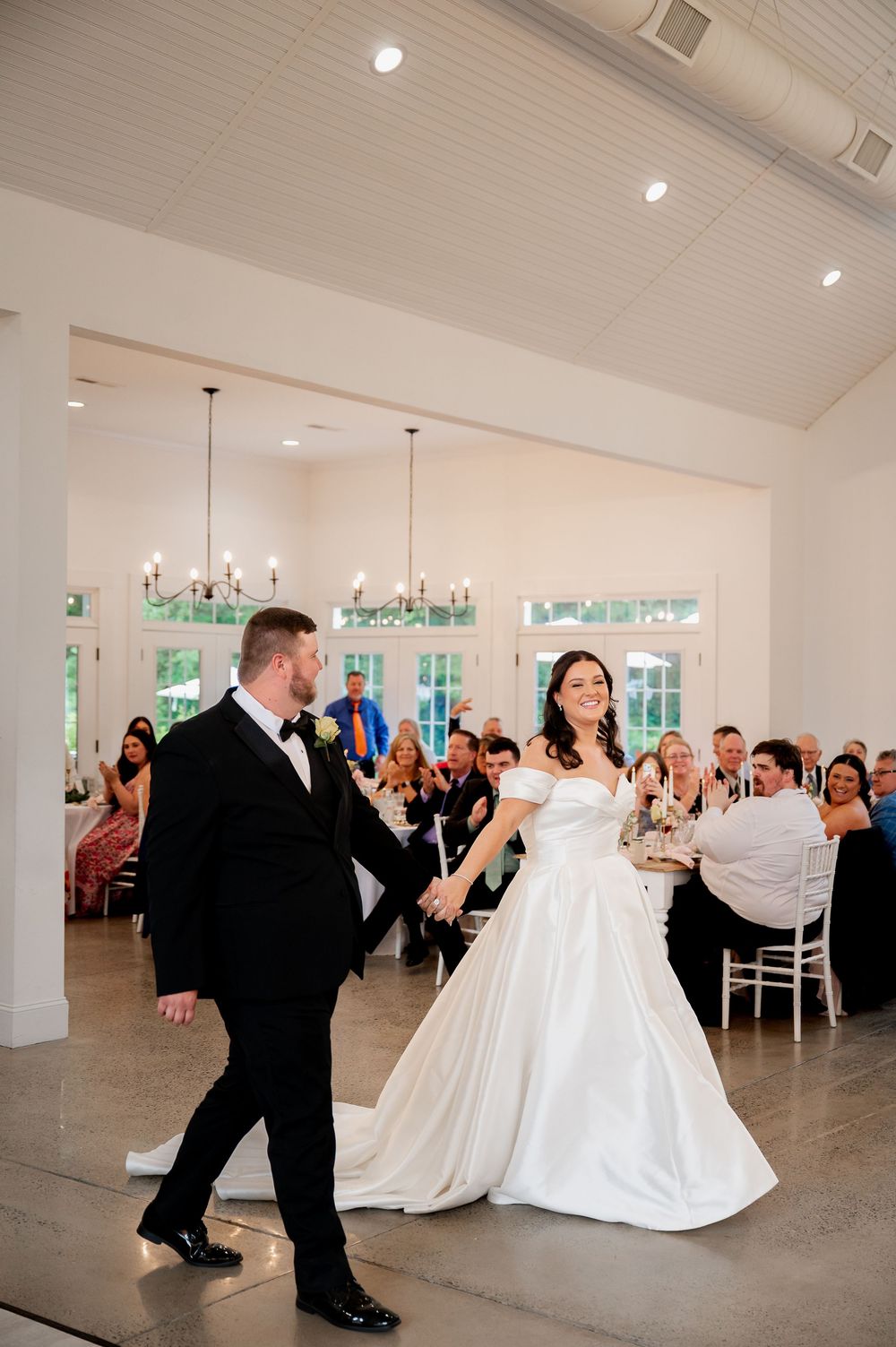 A wedding couple performs their first dance in an elegant white reception venue with chandelier lighting.