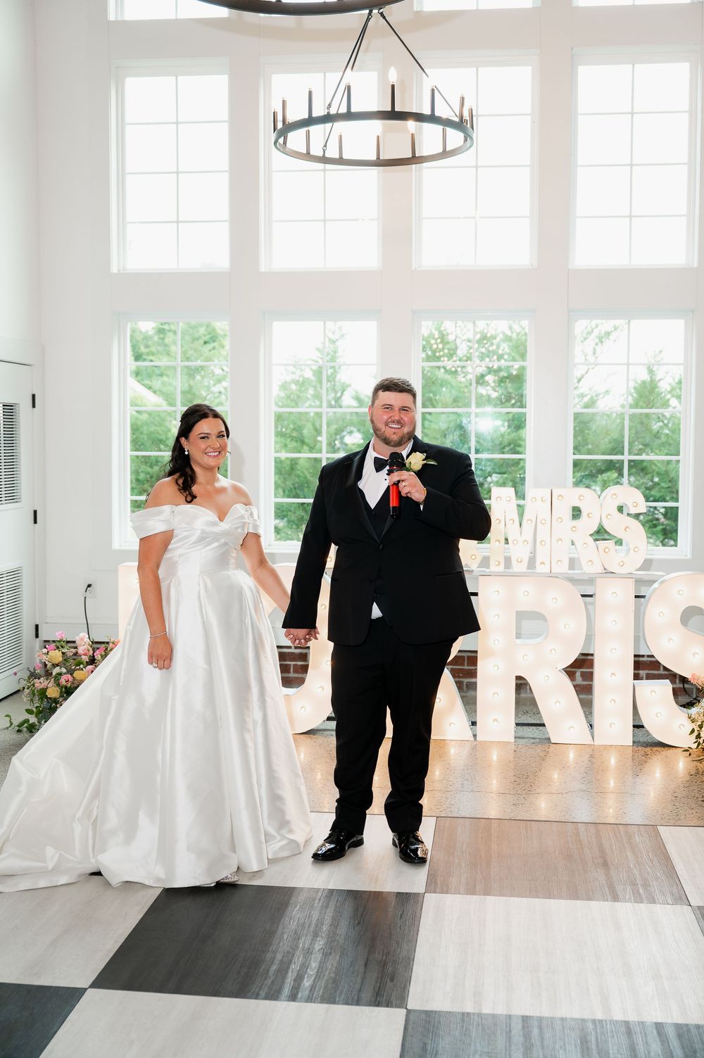 A wedding couple poses in front of illuminated letters in an elegant white venue with checkerboard flooring.