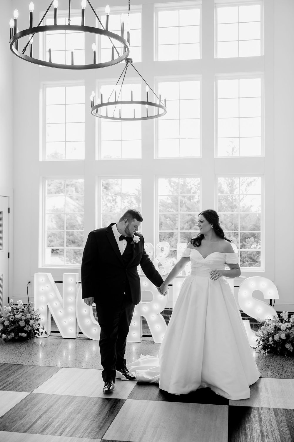 A romantic wedding moment in a bright white venue with dramatic circular chandeliers and tall windows.