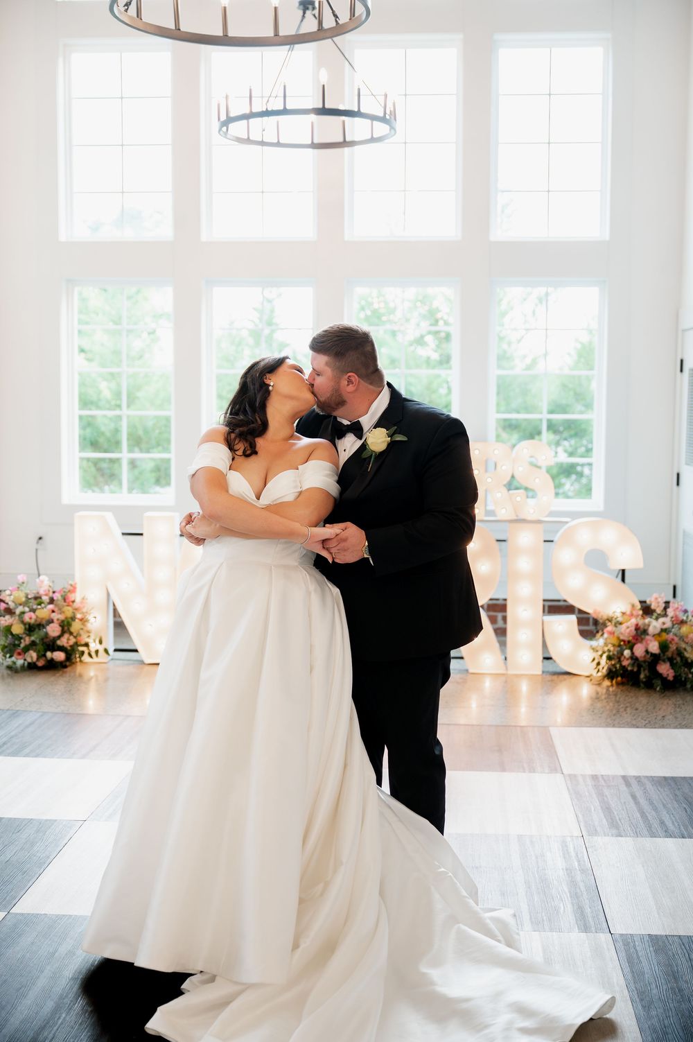 A tender wedding moment captured in an elegant white venue with circular chandeliers and illuminated letters.