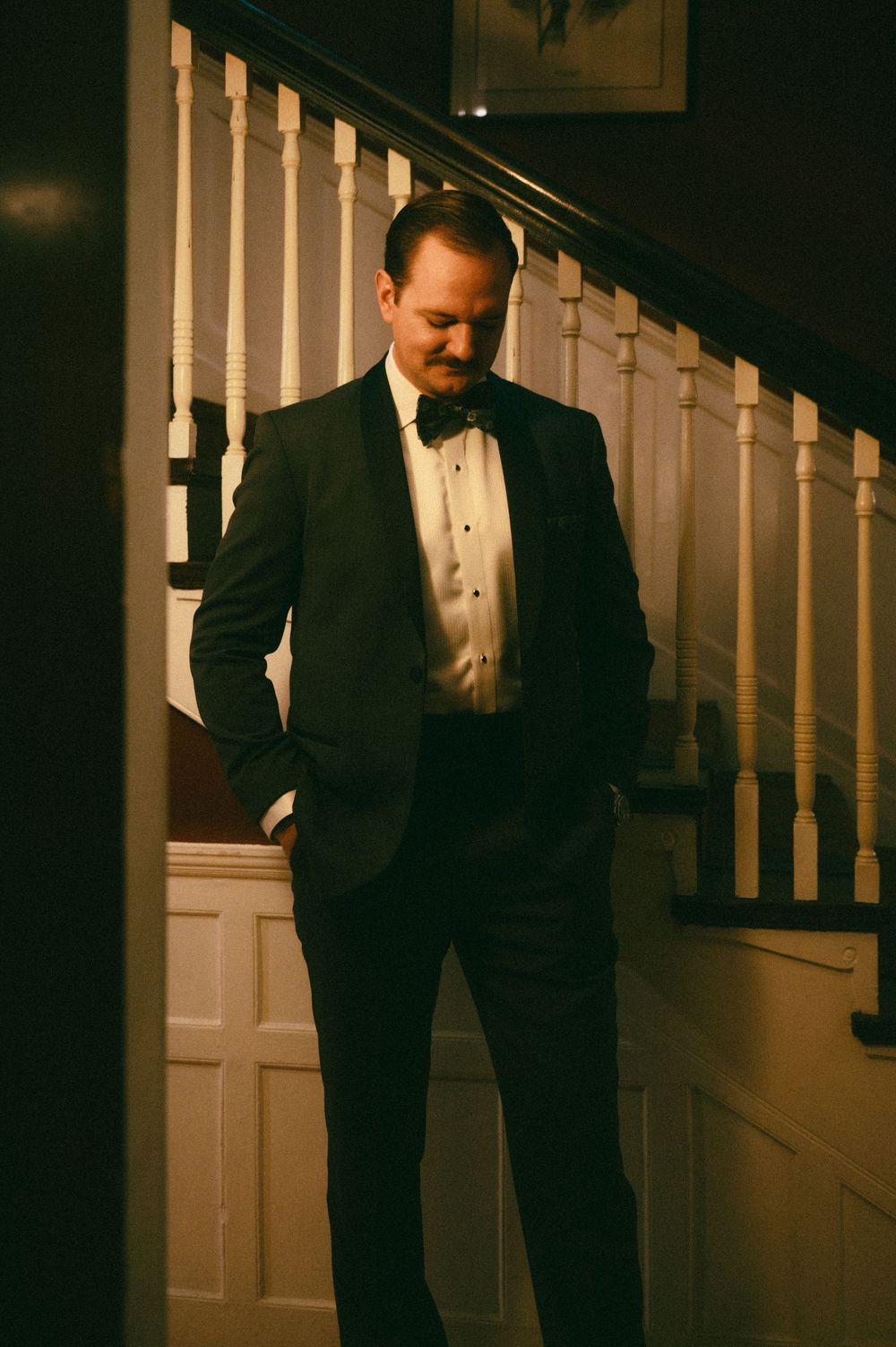 A person in a formal black tuxedo stands on wooden stairs in a dimly lit interior setting.