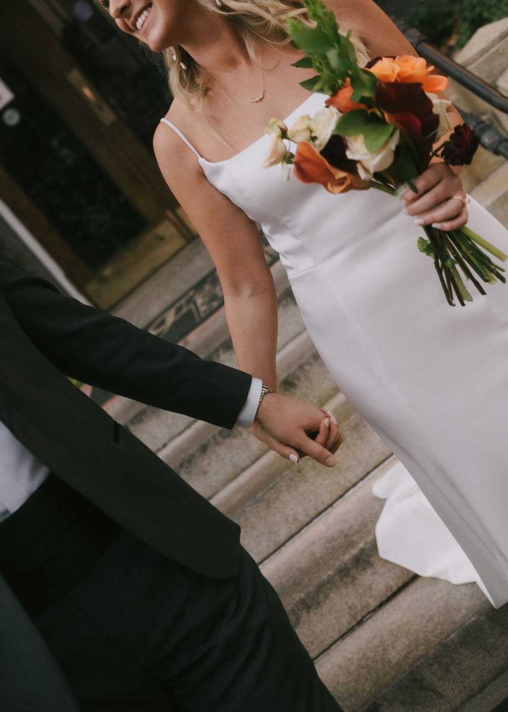 Wedding couple stands on stone steps outdoors with bride holding a vibrant floral bouquet.