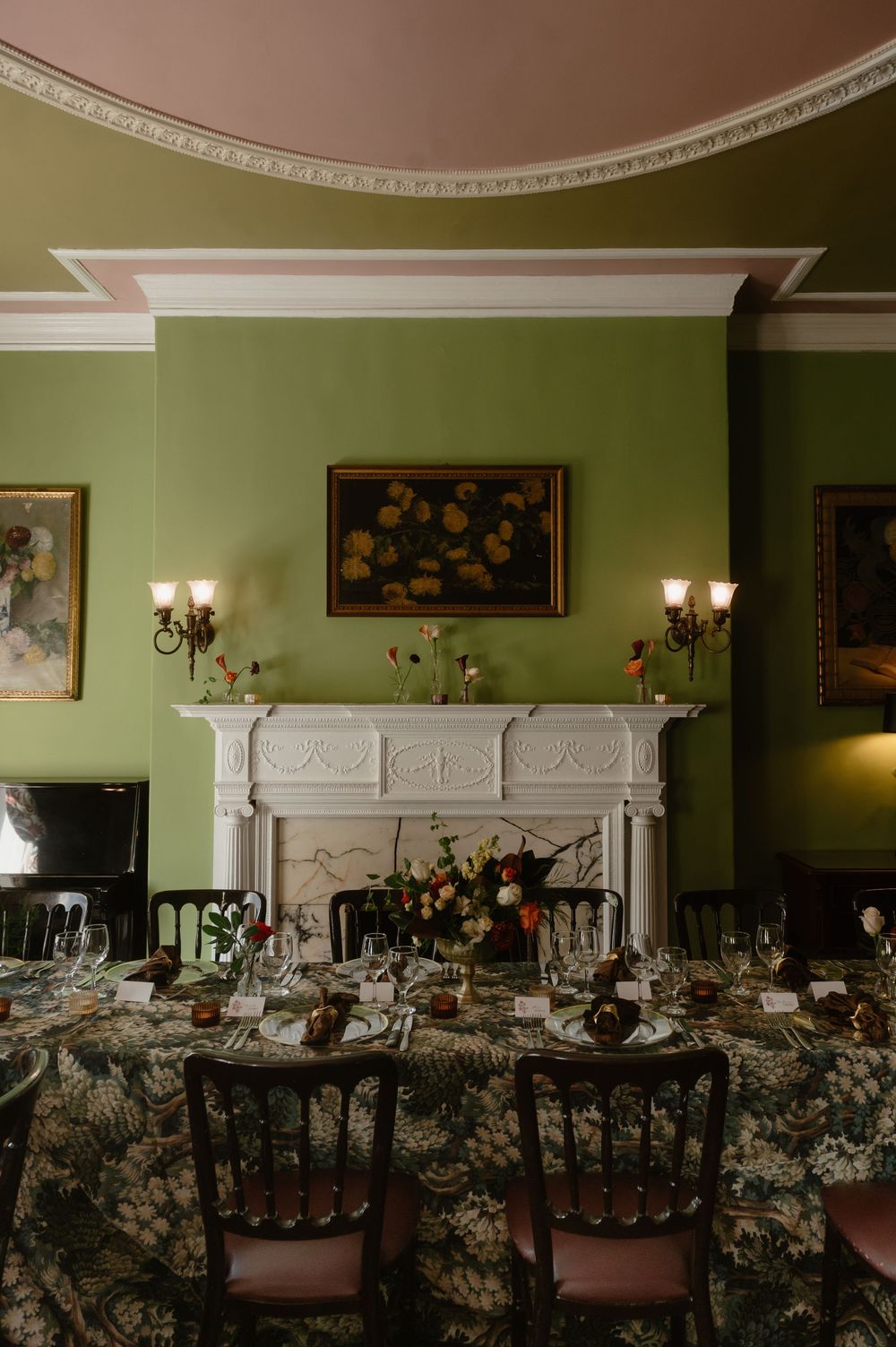 Formal dining room with green walls, ornate white fireplace, and floral table settings.