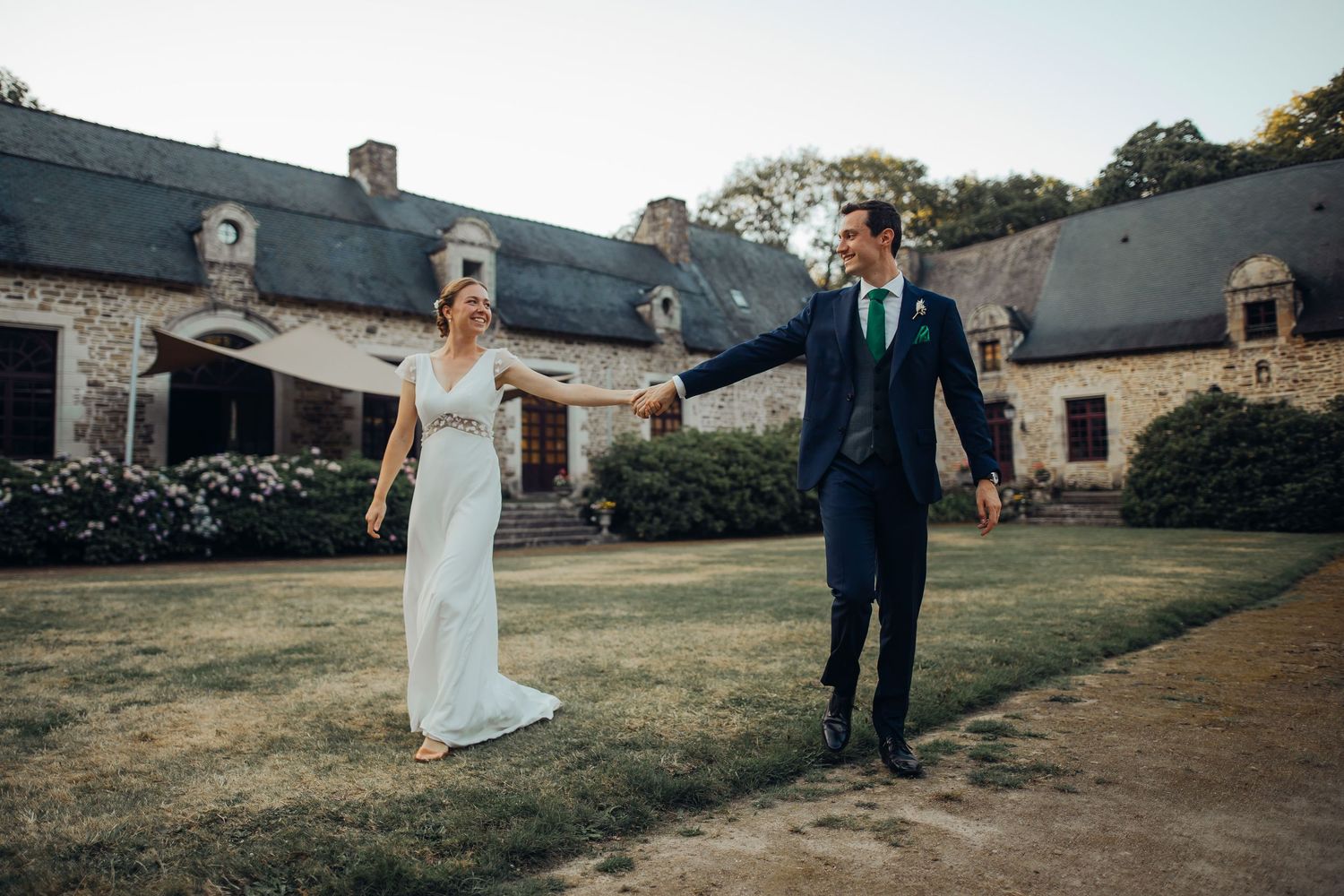 Joyful bride and groom walk hand in hand across lawn before a rustic stone French manor house.