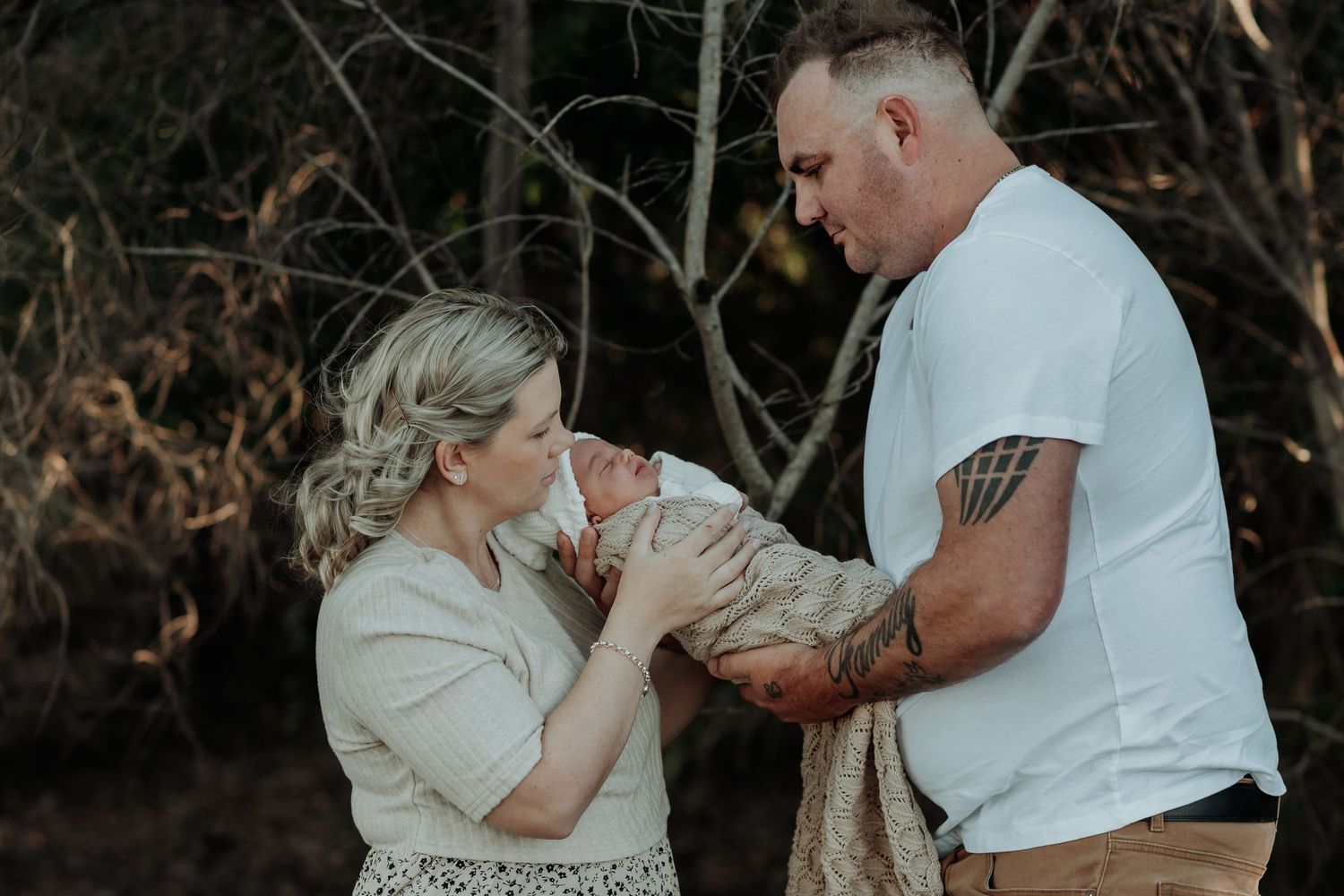 A couple in casual attire share a tender moment outdoors with their newborn baby at sunset.