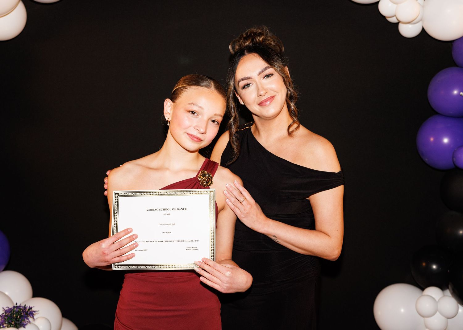 Two people in matching burgundy and black formal dresses pose with a certificate against a dark backdrop with balloons.