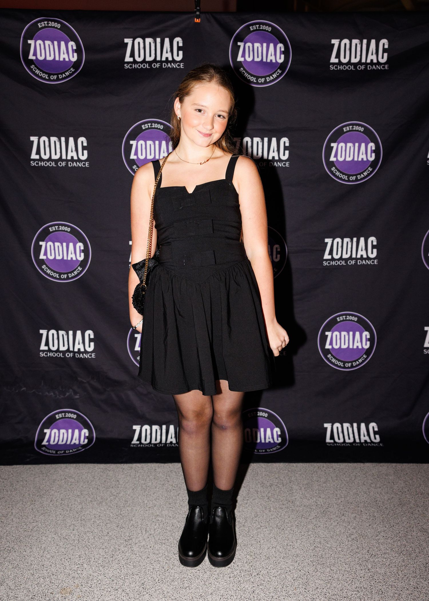 A person in a black dress and boots poses against a purple and black step-and-repeat backdrop with Zodiac logos.