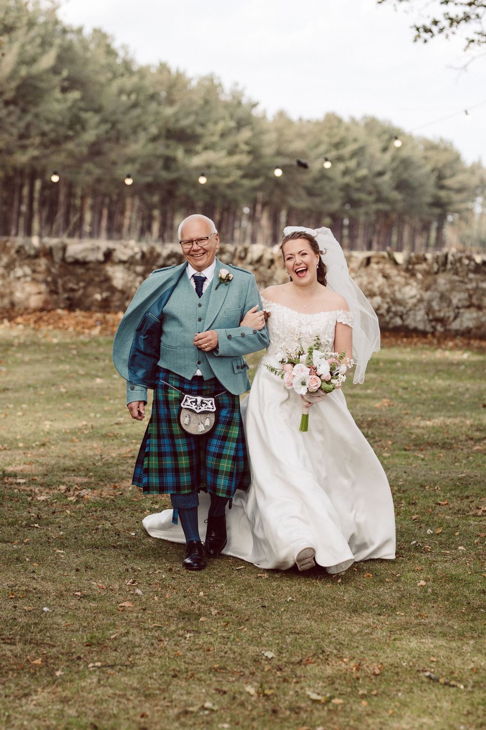 A joyful wedding moment captured as bride walks with companion in traditional Scottish kilt on grassy field.